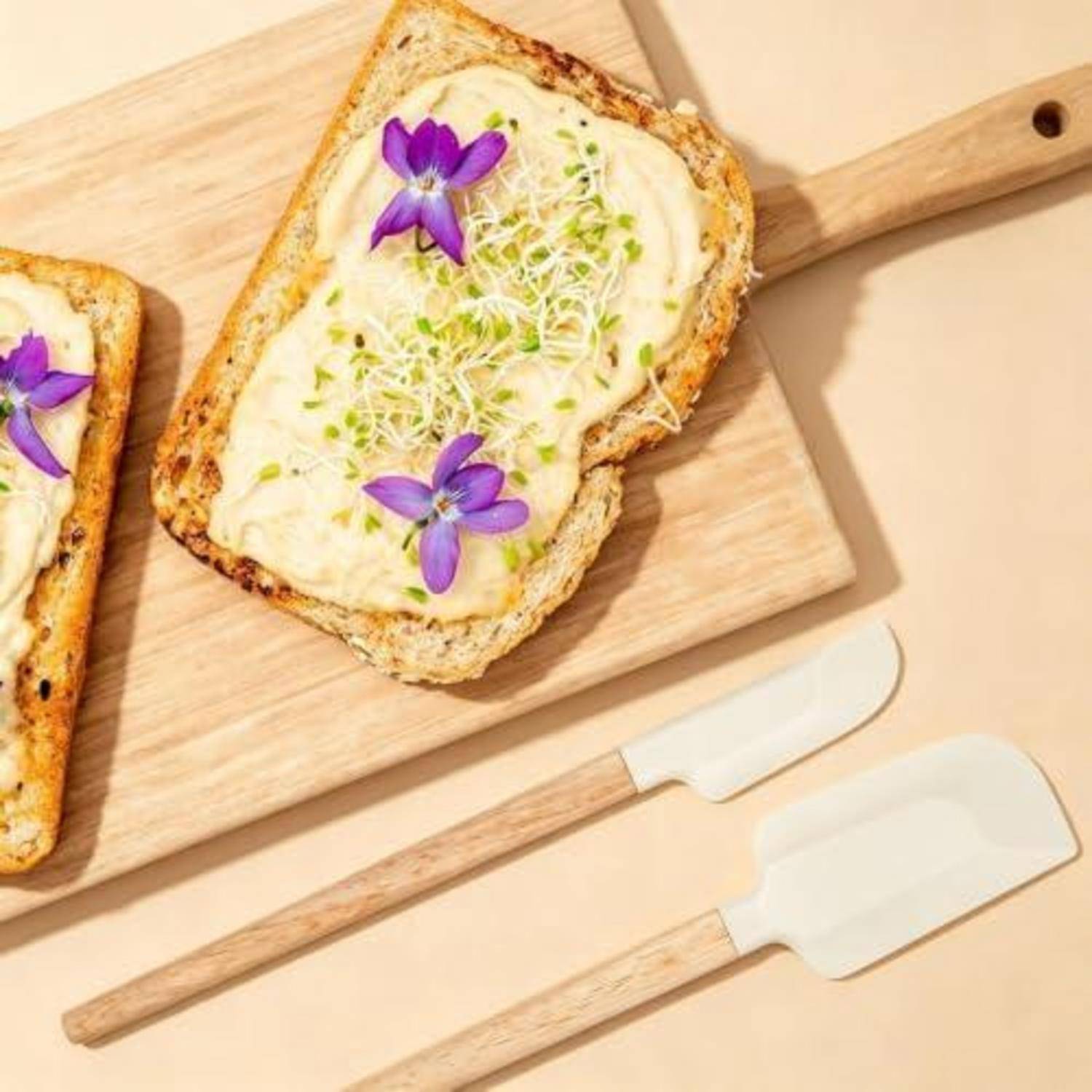 Ein Stück Toast mit cremigem Aufstrich, Sprossen und violetten essbaren Blüten auf einem Schneidebrett belegt, mit zwei Spachteln darunter.