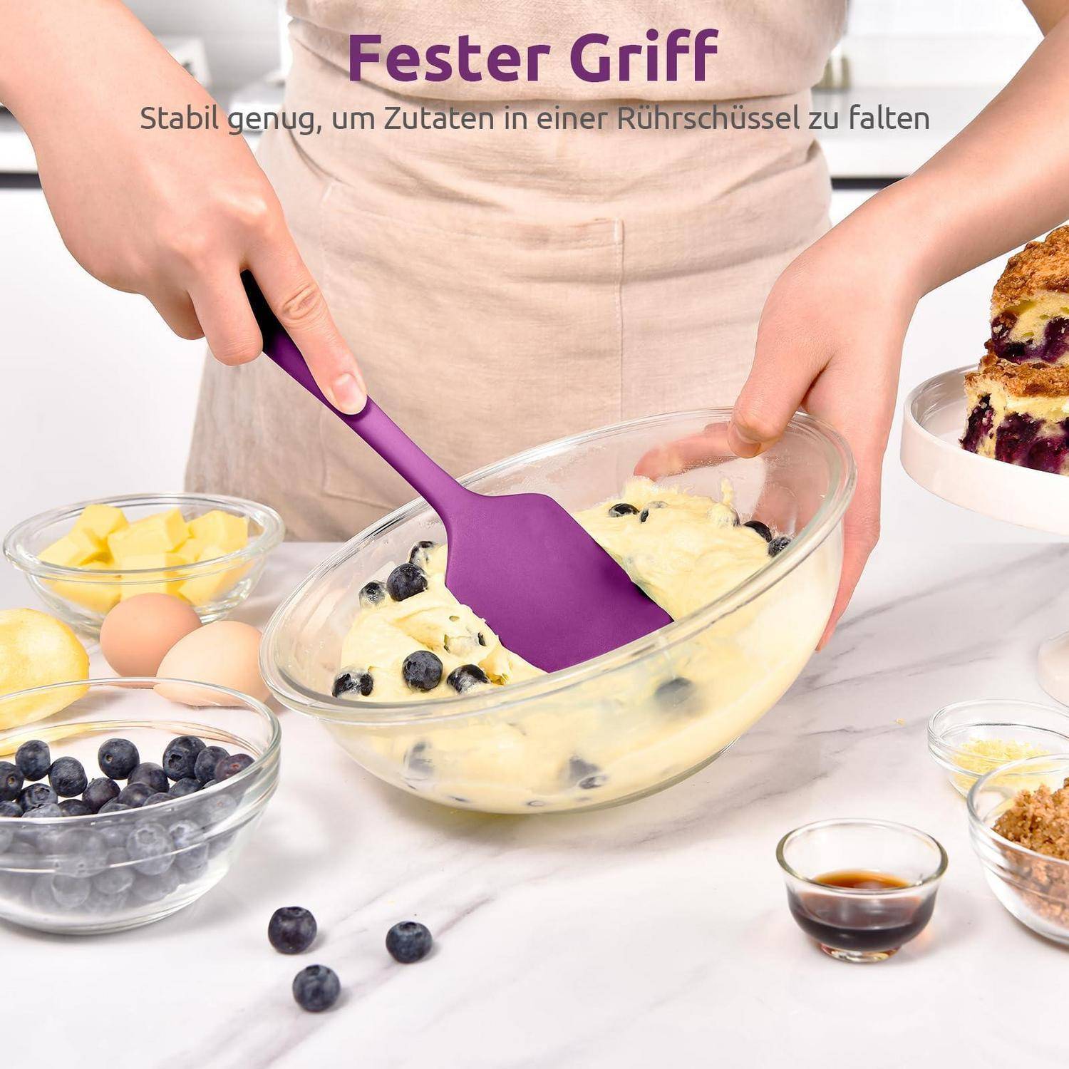 Person, die einen violetten Spatel mit einem 'Ester Griff' verwendet, um Blaubeeren in einen Teig in einer Glassschüssel zu falten, mit Zutaten auf einer Arbeitsplatte.