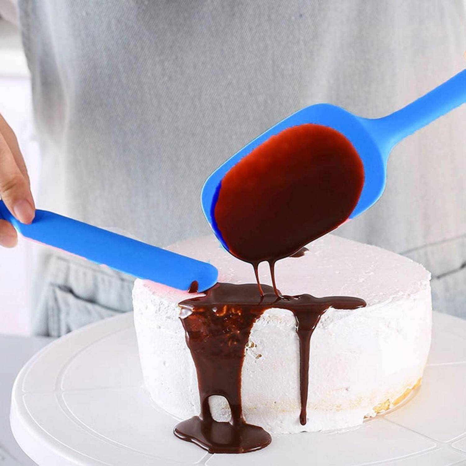 Eine Person verteilt Schokoladensauce mit zwei blauen Spachteln auf einem weiß glasierten Kuchen.