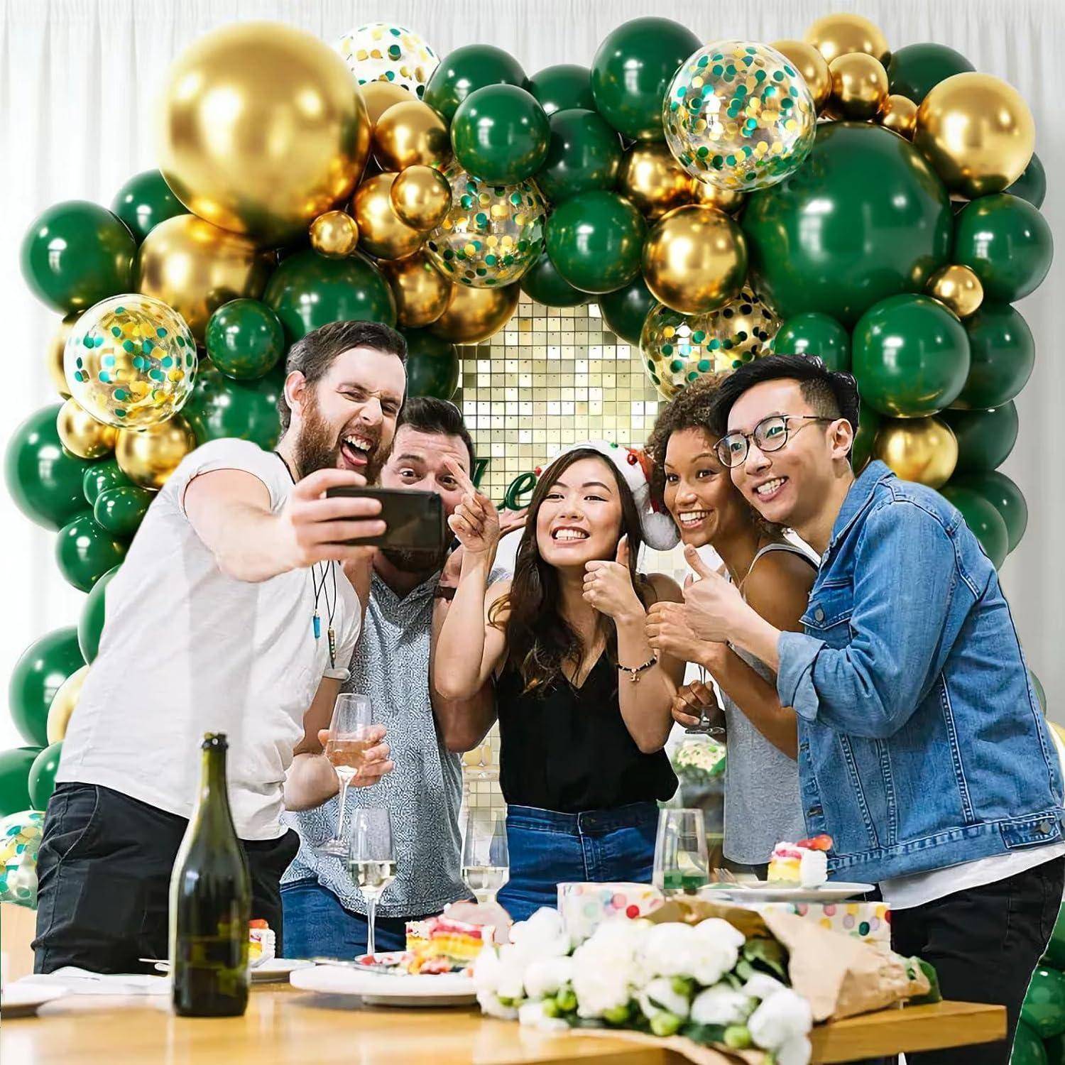 Fünf Freunde posieren für ein Selfie bei einer Feier, lächeln vor einem grün-goldenen Ballonbogen mit einem Tisch voller Kuchen und Getränke.
