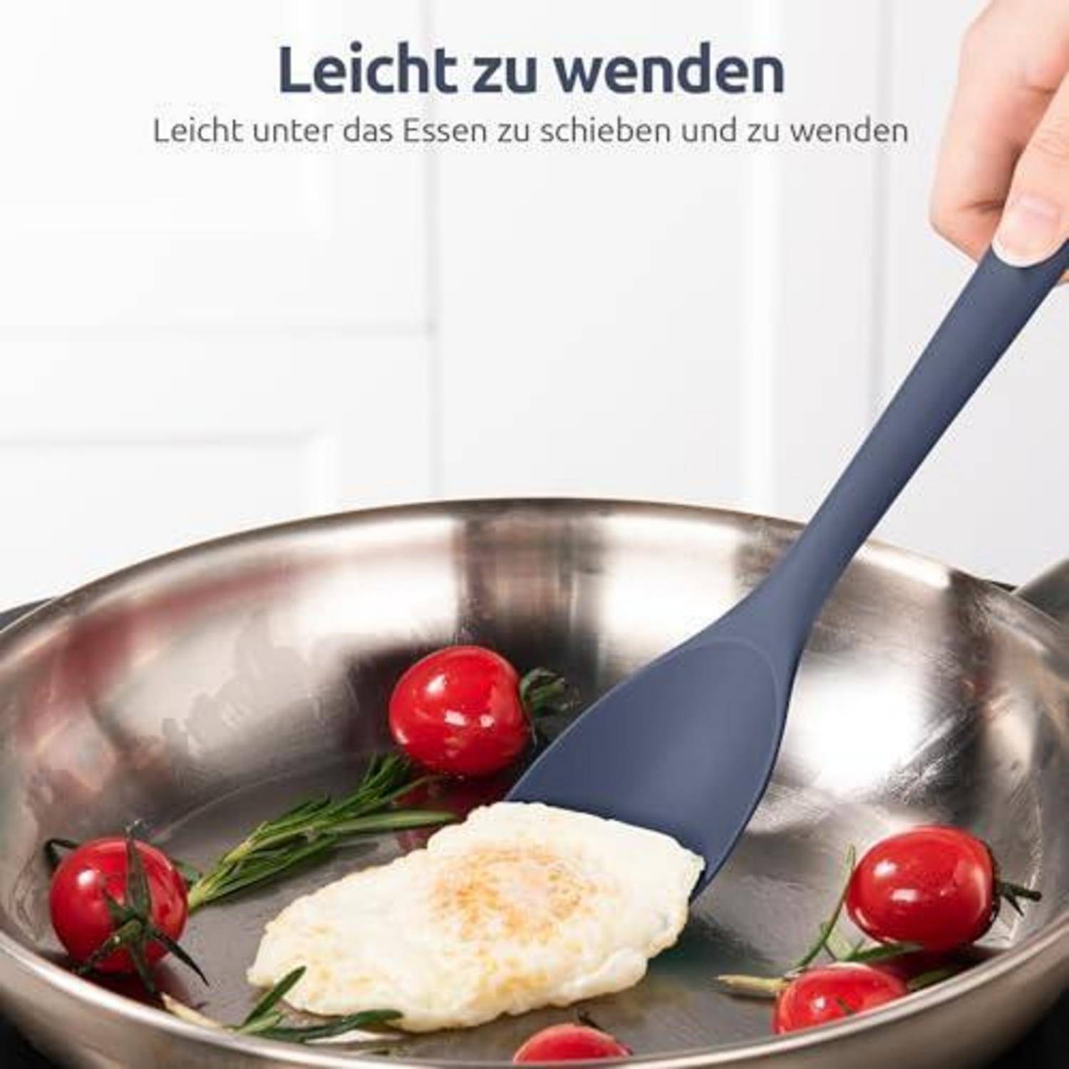 Ein Silikonspatel hebt ein Ei in einer Edelstahlpfanne mit Kirschtomaten; deutscher Text sagt 