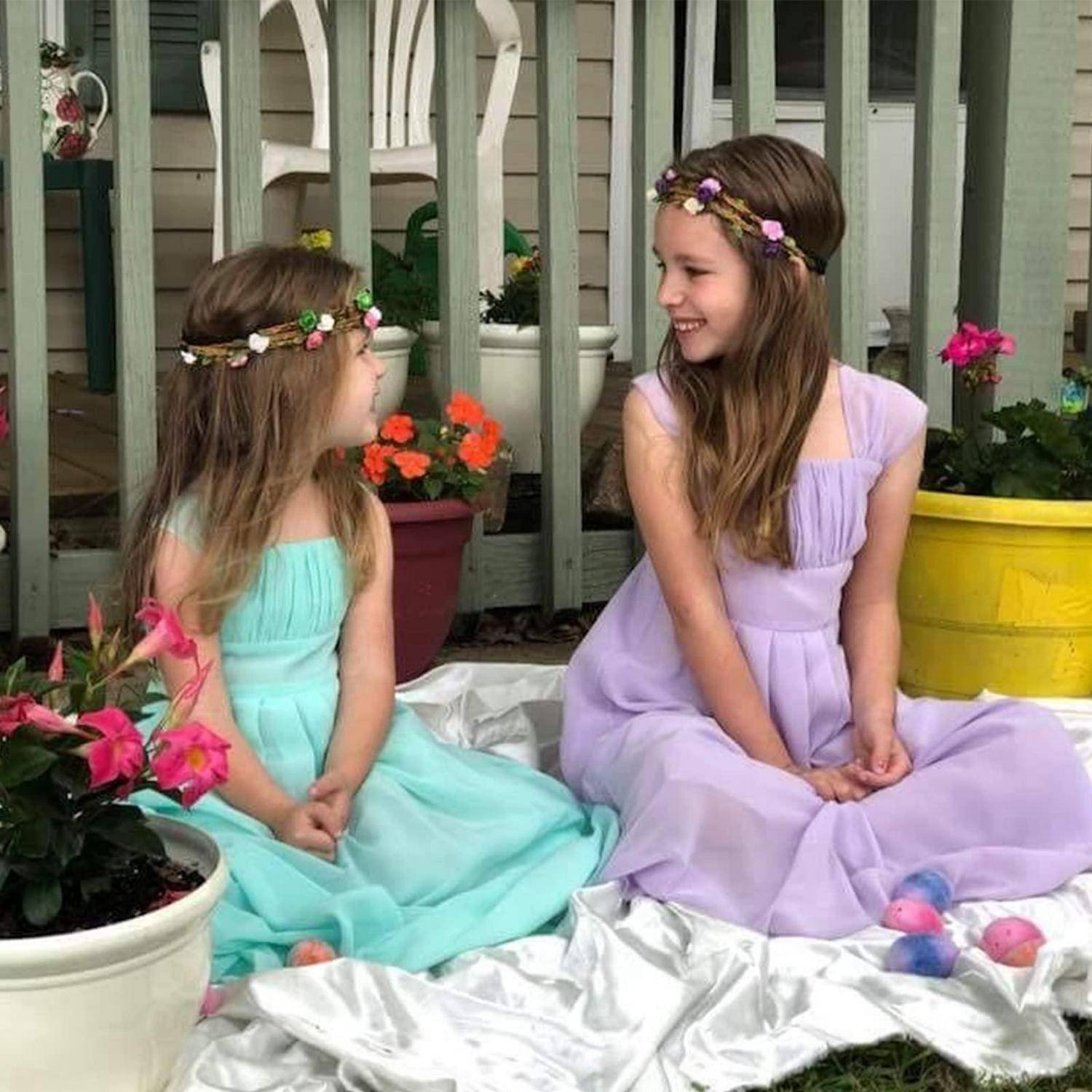 Zwei Mädchen in Pastellkleidern und Blumenkronen sitzen auf einer weißen Decke und lächeln sich in einer gartenähnlichen Veranda-Umgebung an.