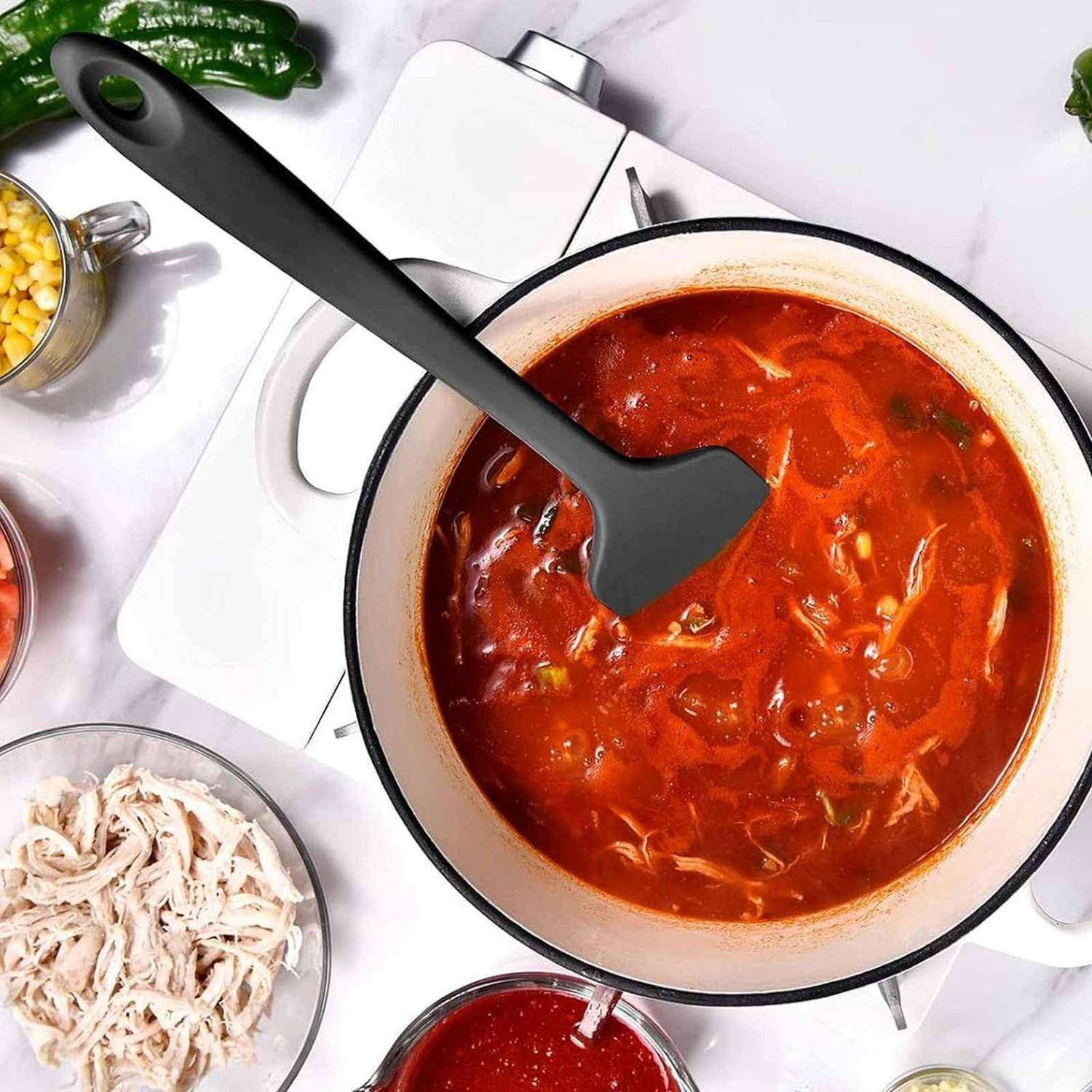 Draufsicht auf einen Topf mit roter Suppe oder Eintopf, der mit einem schwarzen Spatel umgerührt wird, umgeben von zerkleinertem Huhn und Zutaten auf einer Arbeitsplatte.