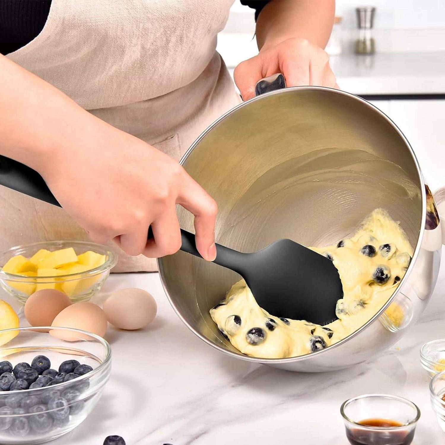 Hände, die Heidelbeer-Teig in einer Metallschüssel mit einem schwarzen Spatel mischen, mit Eiern, Butter und Beeren auf einer Küchenarbeitsplatte.