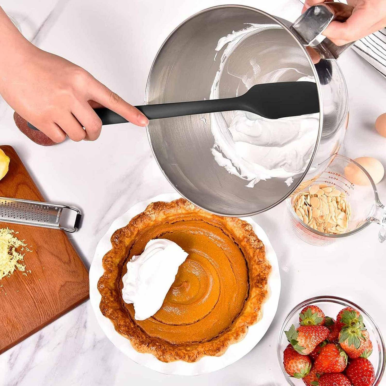 Hände fügen Schlagsahne auf einen Kürbiskuchen auf der Küchenarbeitsplatte hinzu, mit Erdbeeren, Mandeln und Backwerkzeugen in der Nähe.