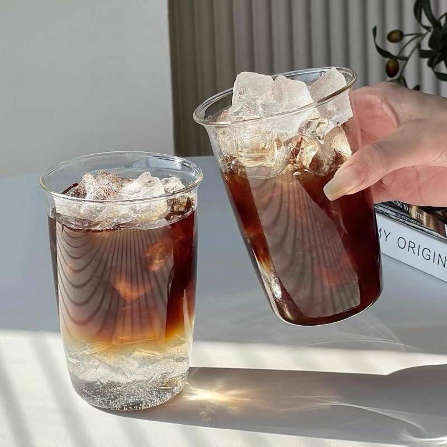 Zwei Eiskaffee-Getränke in durchsichtigen Plastikbechern auf einem Tisch, eines in einer Hand gehalten, mit sichtbarem Eis und dunkel-zu-hell geschichttem Kaffee.