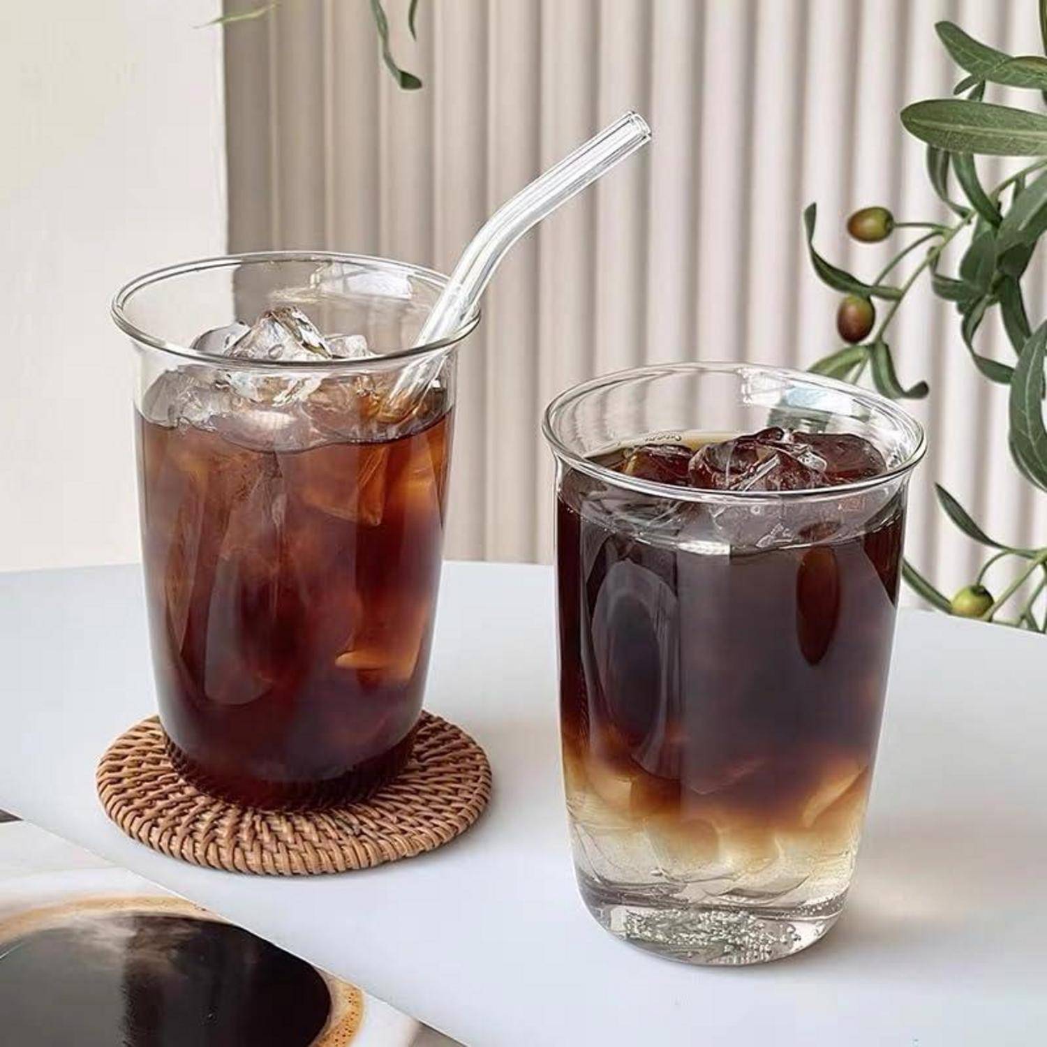 Zwei Gläser Eiskaffee auf einem weißen Tisch, eines mit einem Glasstrohhalm und einem geflochtenen Untersetzer, in einer hellen Innenraumgestaltung.