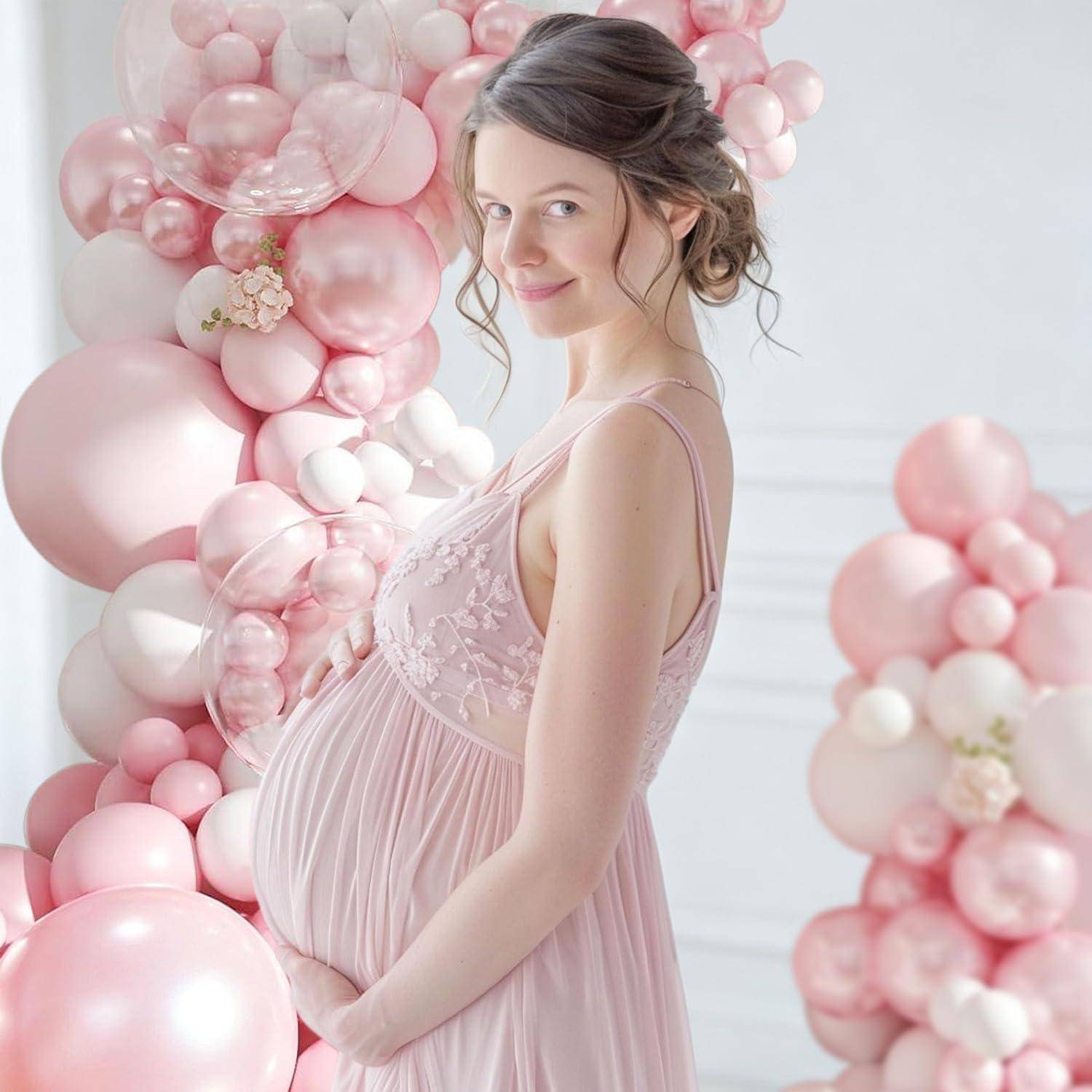Schwangere Frau in einem rosa Kleid, die sanft ihren Bauch hält, neben einer rosa Ballon-Blumen-Kulisse stehend.
