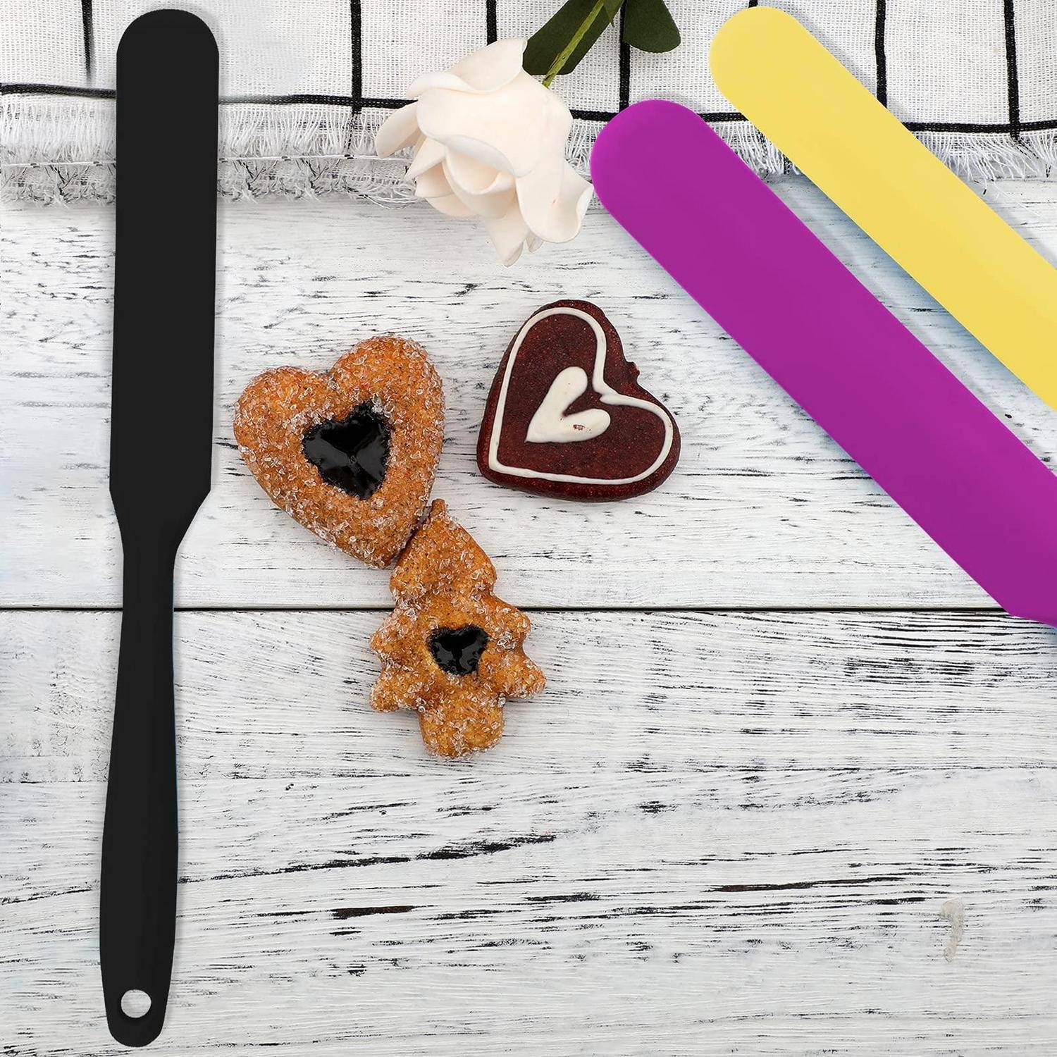 Herzförmige Gebäckstücke mit Marmelade, plus ein schwarzer Spatel und bunte Utensilien auf einem weißen Holztisch mit Stoffhintergrund.