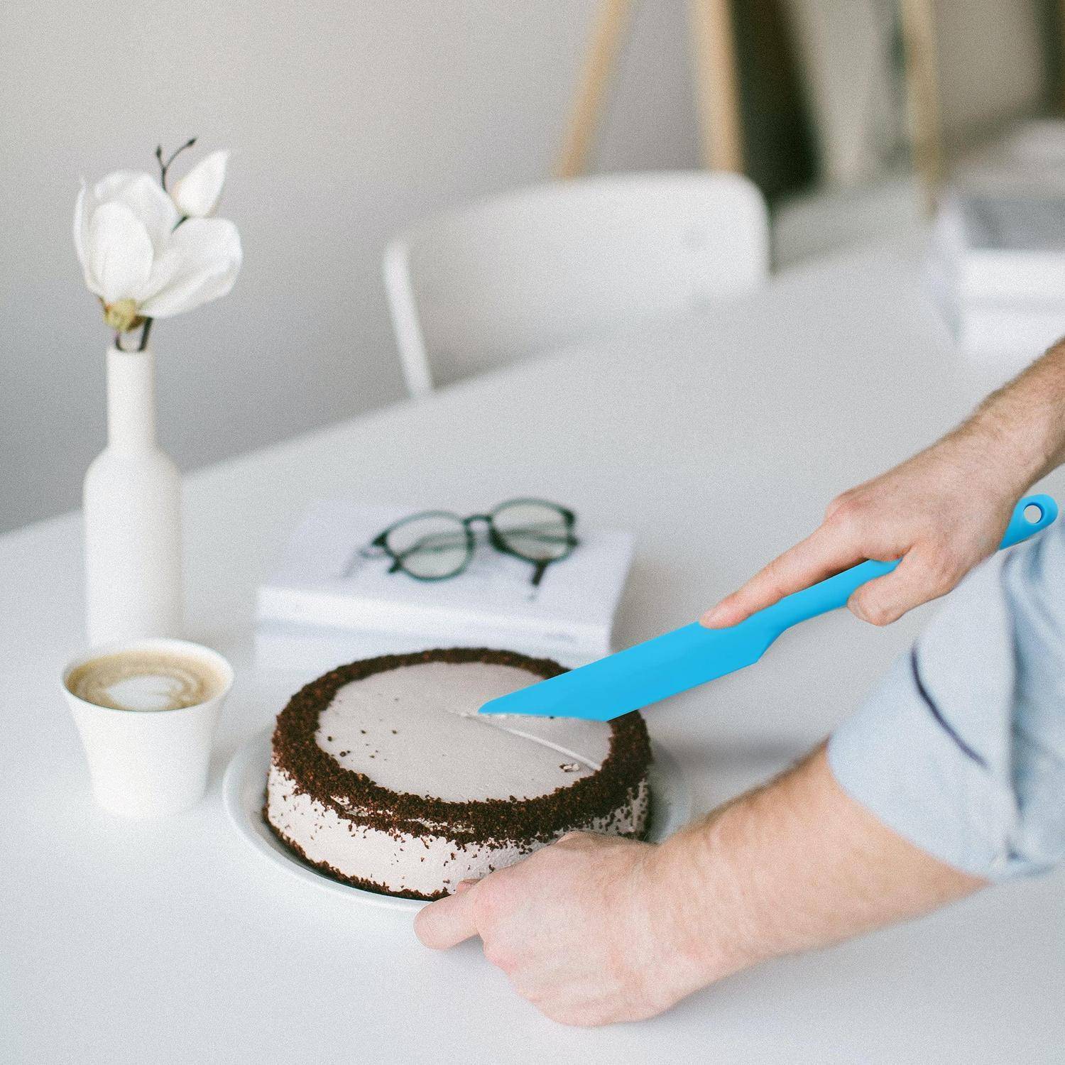 Eine Person schneidet einen glasierten Kuchen auf einem weißen Tisch neben einer Tasse Kaffee, einer Brille und einer Vase mit Blumen.