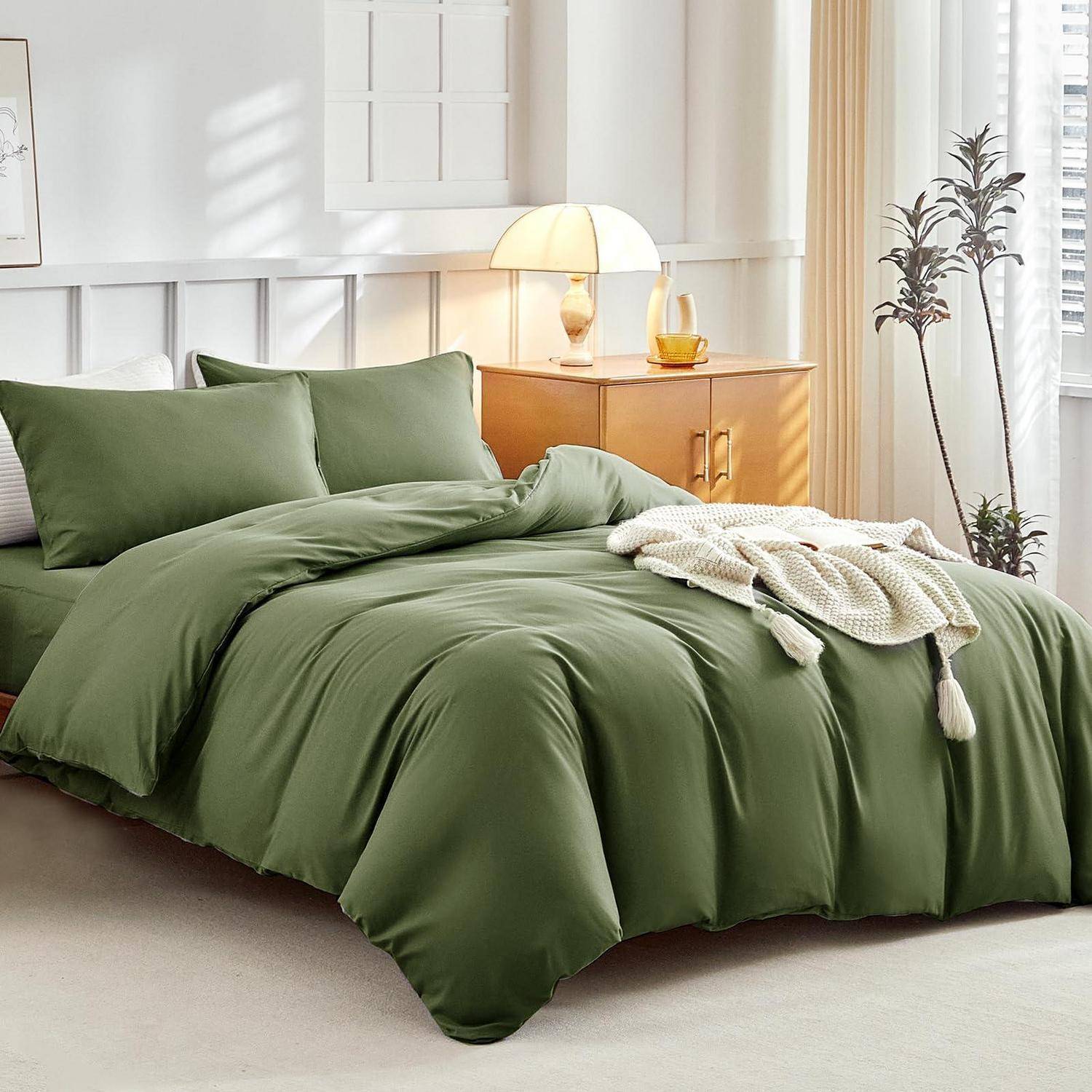 Ein ordentlich gemachtes Bett mit olivgrüner Bettwäsche, zwei Kissen, einer weißen Strickdecke und einer Nachttischlampe in einem hellen, minimalistischen Schlafzimmer.