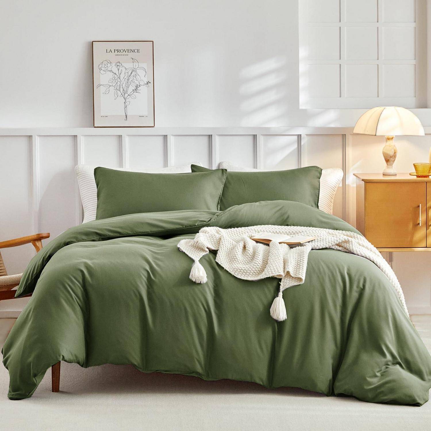 Sorgfältig gemachtes Bett mit olivgrüner Bettwäsche, weißer Wurfdecke, Nachttischlampe und gerahmtem Wandbild in einem hellen, minimalistischen Schlafzimmer.