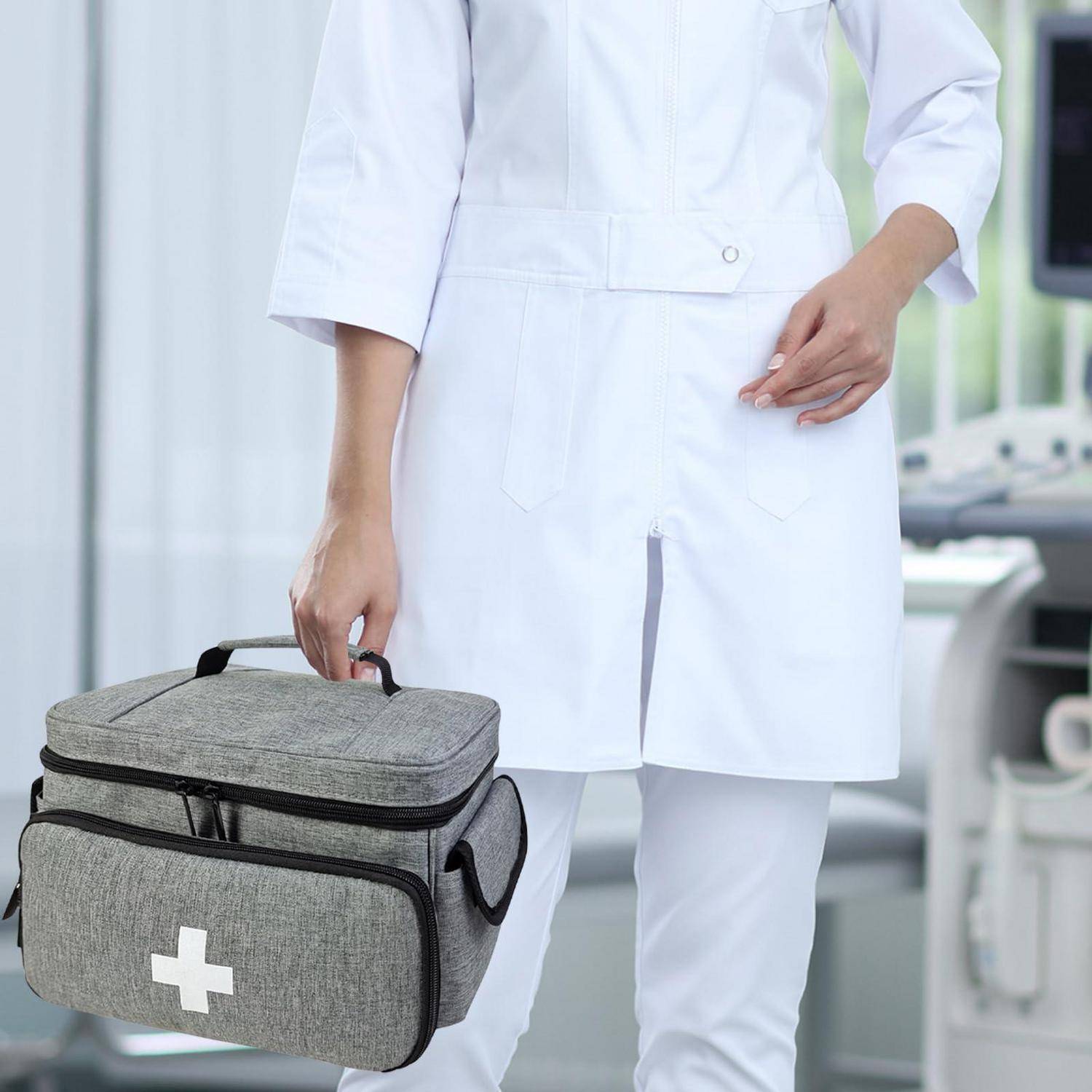 Eine Krankenschwester in weißer Uniform hält eine graue Erste-Hilfe-Tasche mit weißem Kreuz in einer klinischen Umgebung.