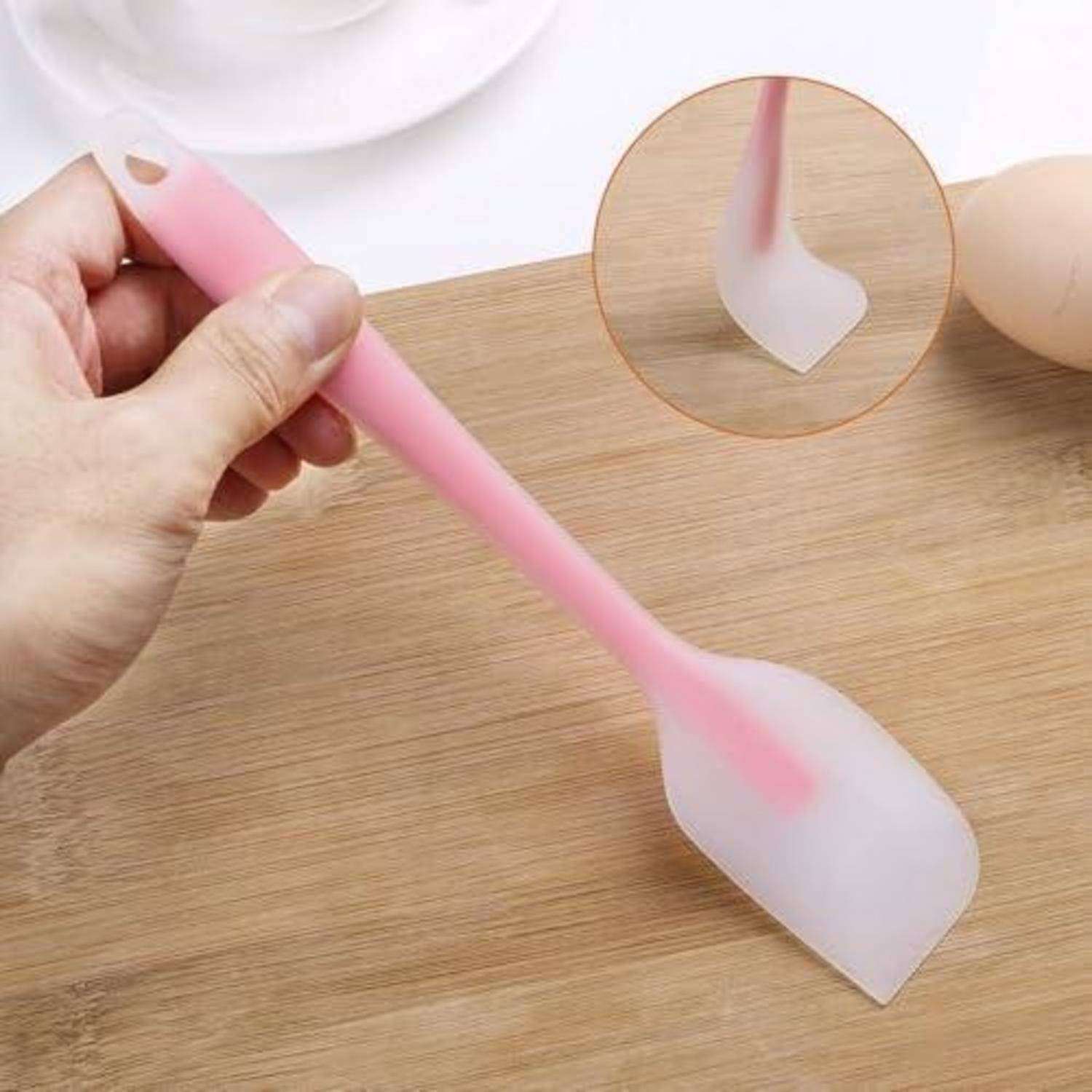 Hand, die einen rosa Silikon-Spatel mit weißem flexiblem Kopf auf einer Holzoberfläche hält, mit einer kreisförmigen Einfügung, die das gekrümmte Spatelende in Detail zeigt.