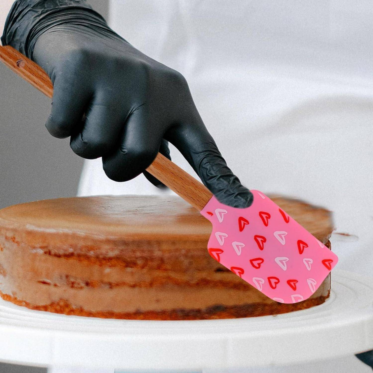 Behandschuhte Hand verwendet einen rosa herzgemusterten Teigschaber, um Zuckerguss auf einem geschichteten Kuchen auf einer Drehplattform zu glätten.