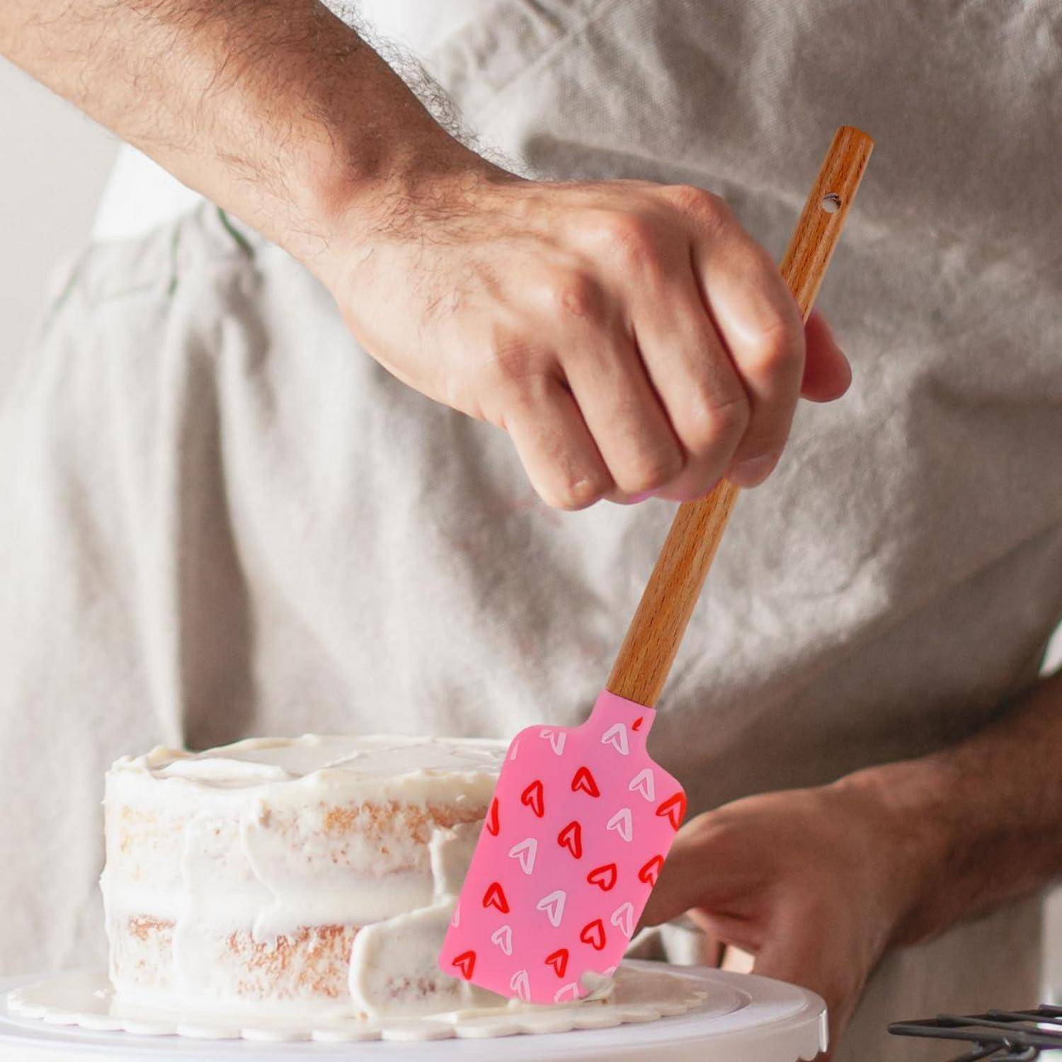 Eine Person, die einen weißen Schichtkuchen mit einem rosafarbenen Spatel mit Herzenmuster auf einem Kuchenständer glasiert.