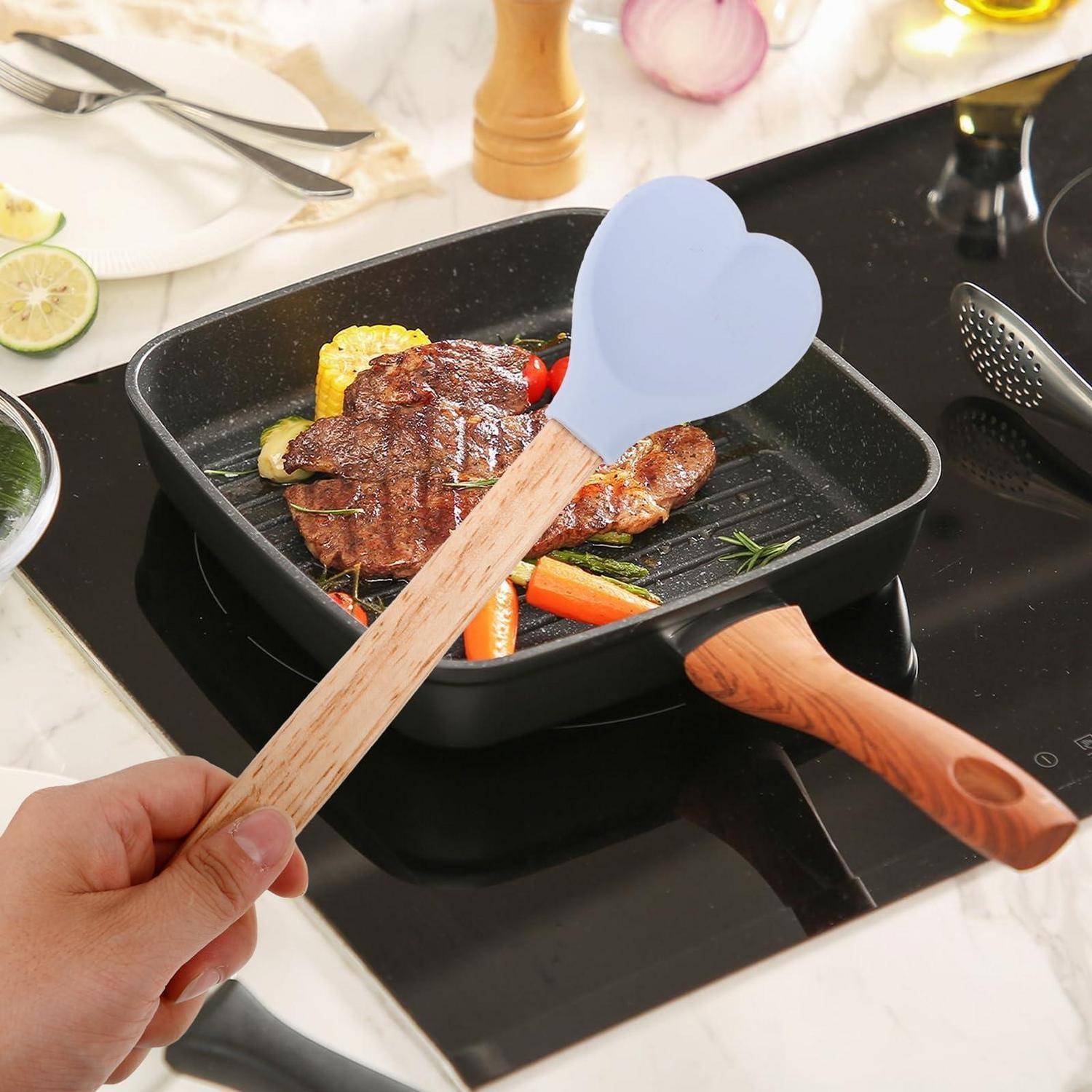 Hand hält einen Holzgriff mit blauem herzförmigem Spatel über einer Grillpfanne mit Steak und Gemüse auf einem Kochfeld.