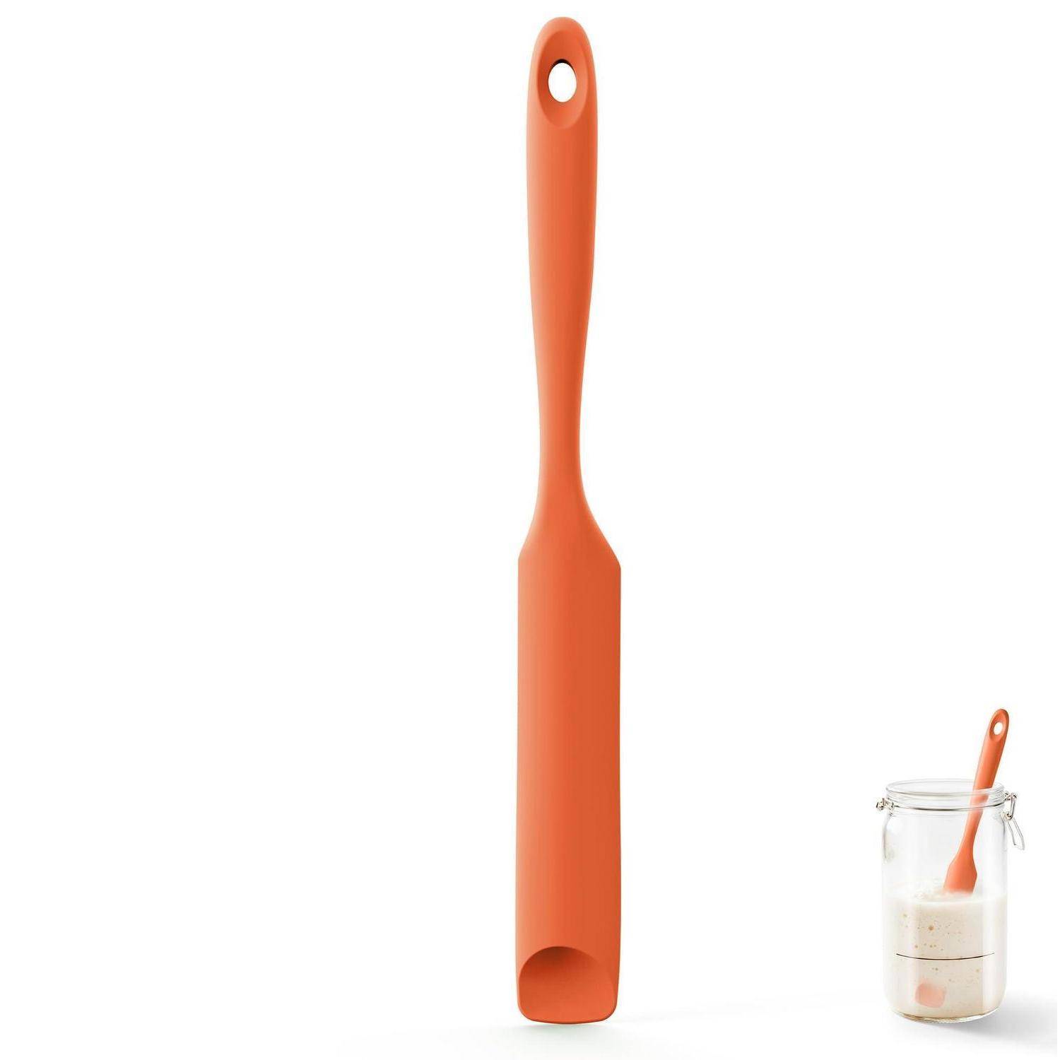 Orange-farbiger Silikonspatel mit Aufhängeloch, aufrecht auf weißem Hintergrund dargestellt, mit einem kleinen Behälter Teig und Löffel unten rechts.