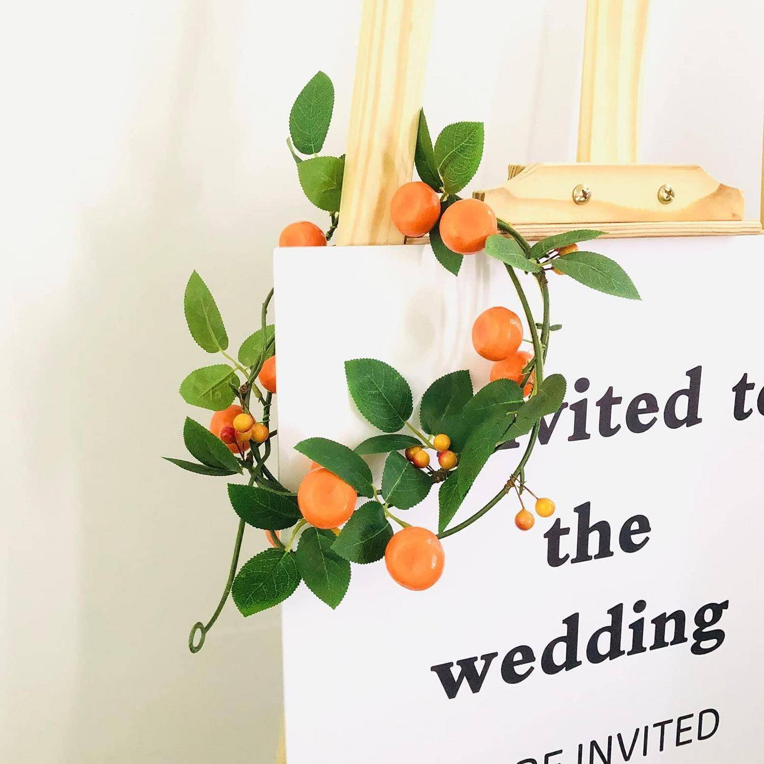 Ein weißes Hochzeitseinladungsschild, das mit grünen Blättern und orange Früchten verziert ist, mit den teilweise sichtbaren Worten „zur Hochzeit eingeladen".