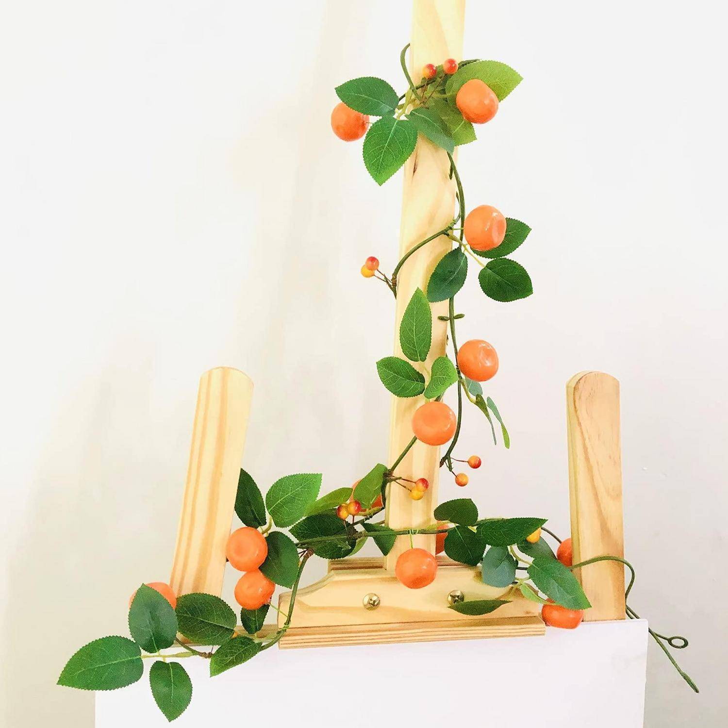 Künstliche orangefarbene Beerenranke, die um einen Holzrahmen gewickelt ist, mit grünen Blättern und orangefarbenen Früchten vor einem einfachen weißen Hintergrund.