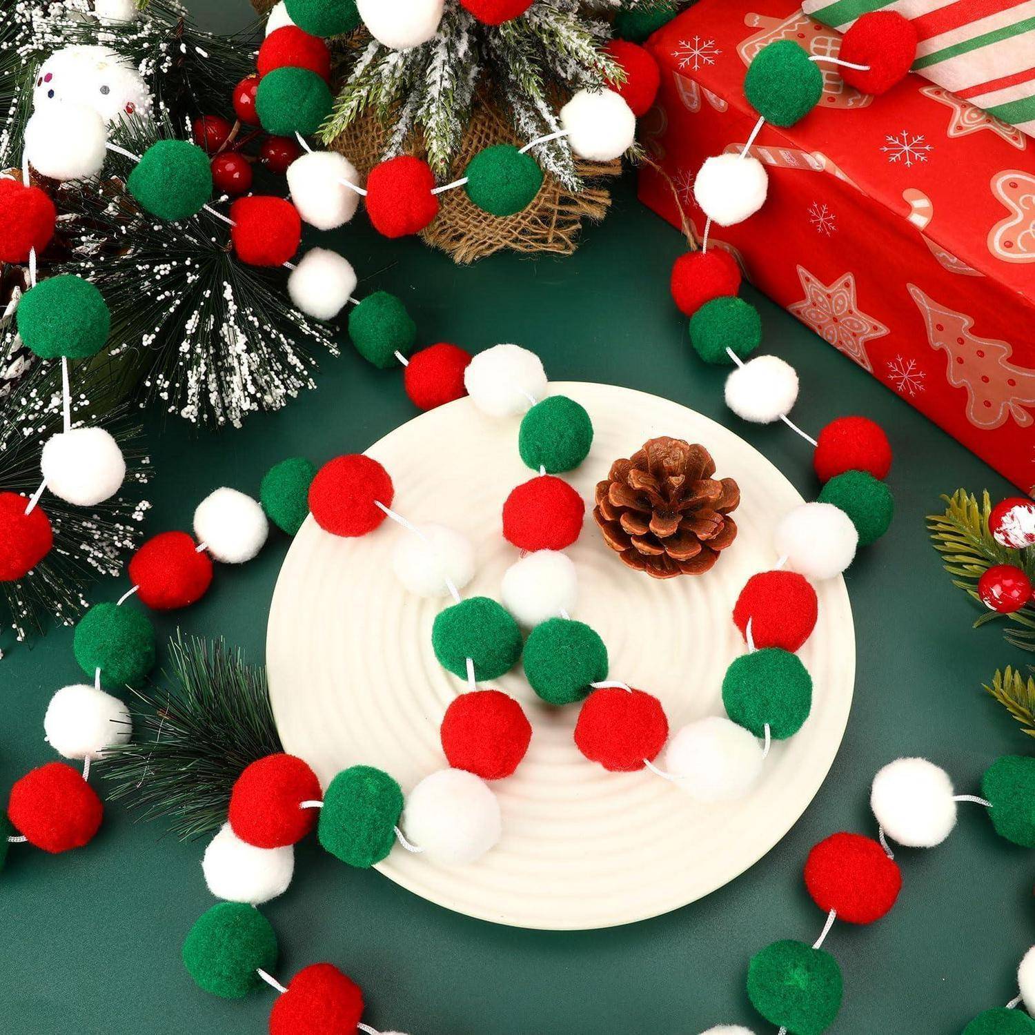 Weihnachtliches Bastelzimmer mit roten, grünen und weißen Bommel-Girlanden um einen weißen Teller, der einen Tannenzapfen hält, mit Geschenkboxen und festlicher Grünflora.