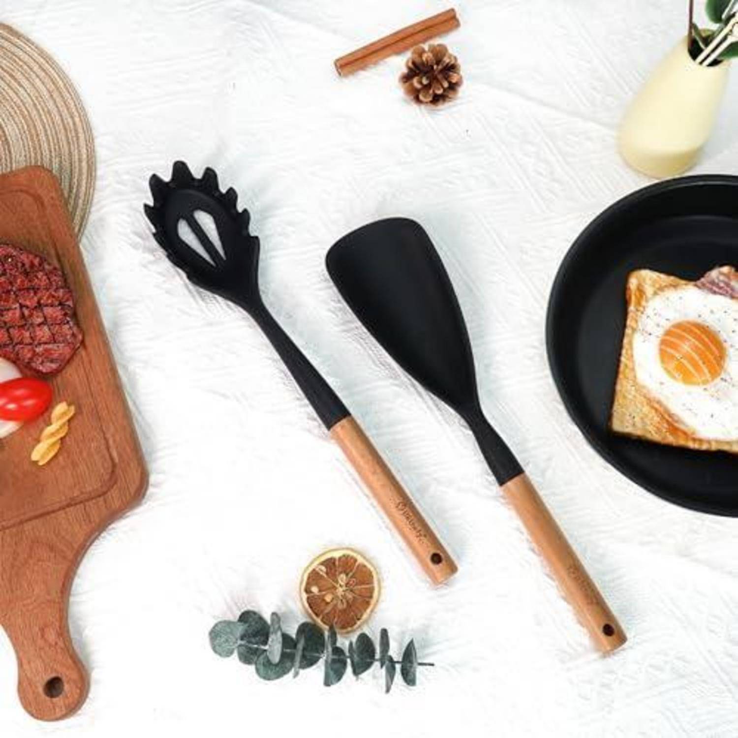Küchenutensilien und Frühstücksgegenstände auf einer weißen Oberfläche, einschließlich zwei schwarz-holzerner Kochlöffel, einer Bratpfanne mit Ei-Toast und einem Schneidebrett mit Fleisch.