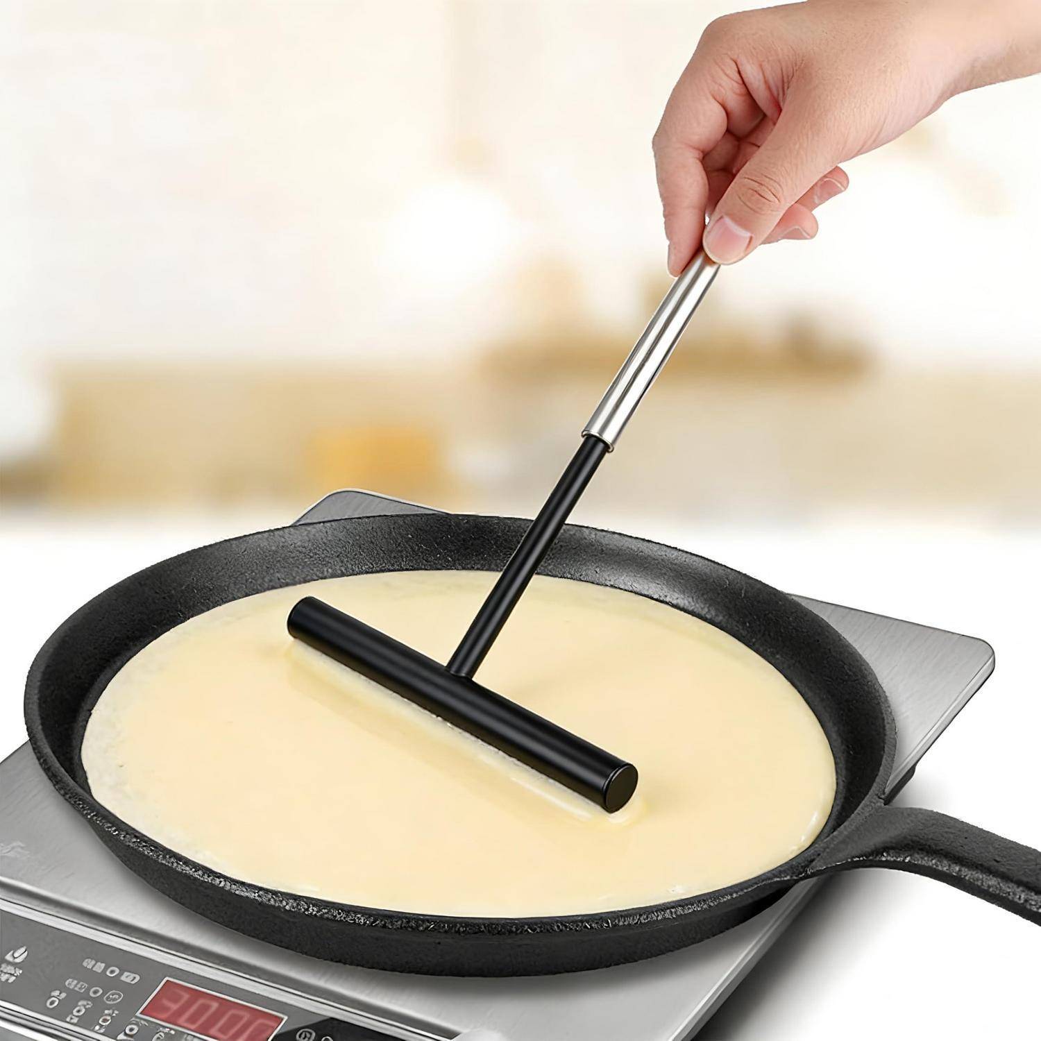 Hand mit einem T-förmigen Teigverteiler, der Crêpe- oder Pfannkuchenteig in einer runden Pfanne auf einem Kochfeld glättet.
