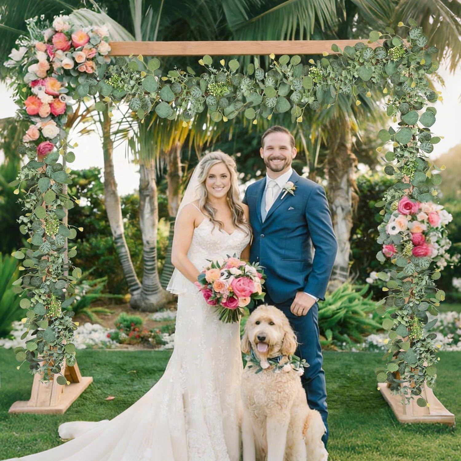 Braut und Bräutigam posieren unter einem blumengeschmückten Hochzeitsbogen mit ihrem Hund in einem tropischen Garten.