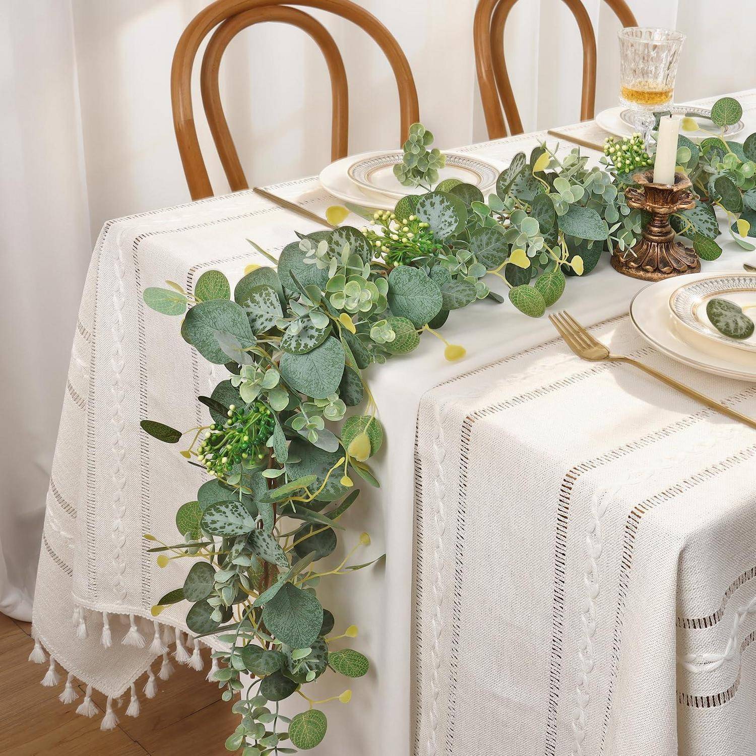 Esstisch mit weißer strukturierter Tischdecke, grüner Eukalyptus-Girlande als Tischdekoration, Holzstühlen und goldfarbig akzentuierten Platzdeckchen