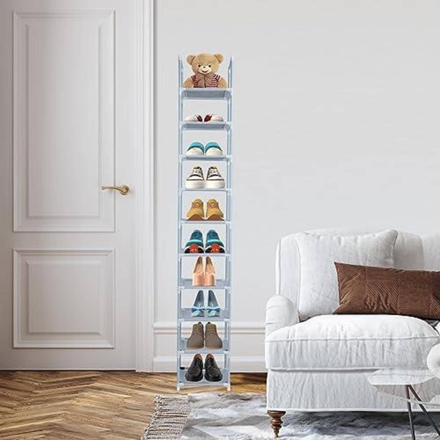 Hohes, transparentes Schuhregal neben einem weißen Sofa und einer Tür, das mehrere Paar Schuhe enthält und auf dem oben ein Teddy sitzt, in einem hellen Raum.