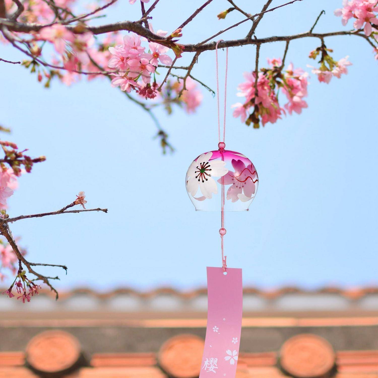Zarte Kirschblüten rahmen ein hängendes gläsernes Windspiel mit einem rosa Papieretikett gegen einen klaren blauen Himmel ein.