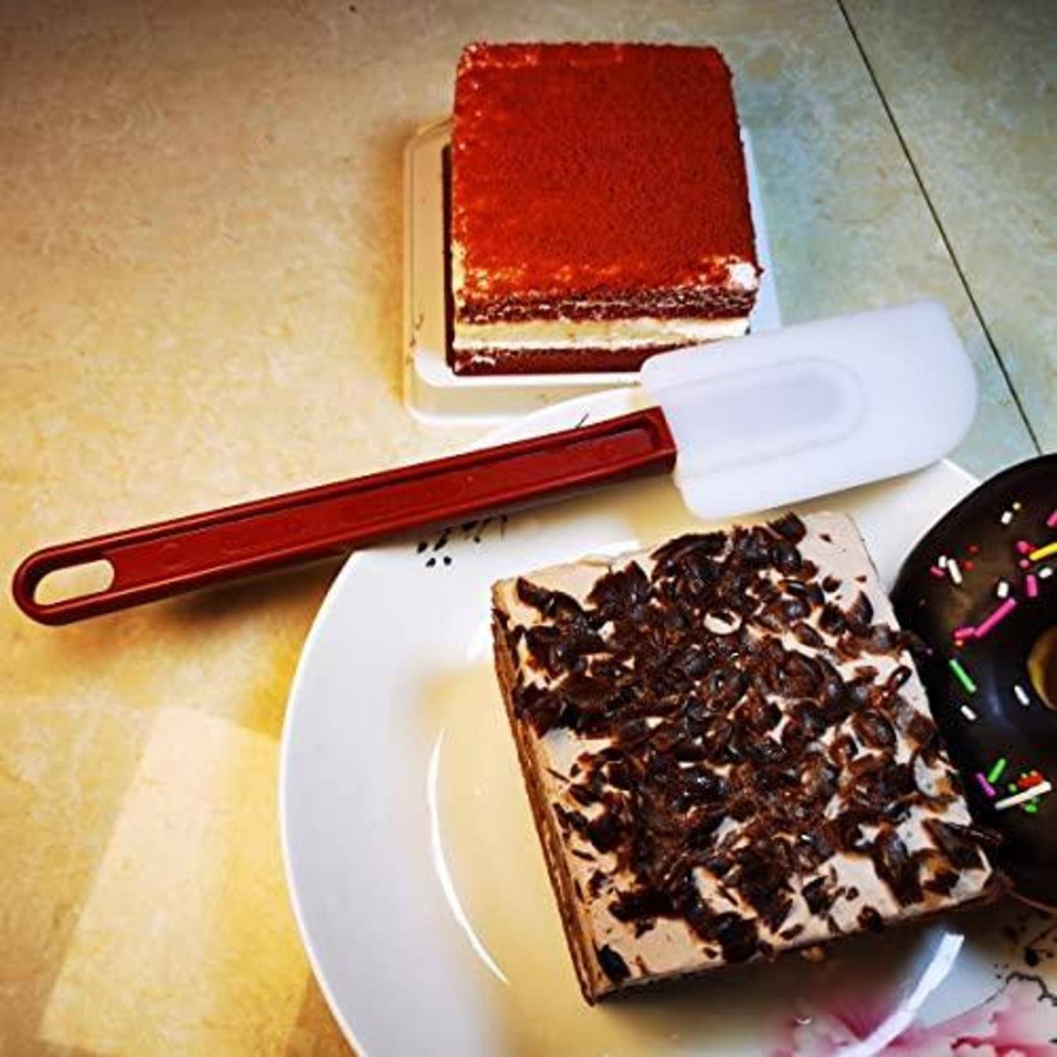 Verschiedene Desserts auf einer Arbeitsplatte: ein Stück Kuchen mit Schokoladenbelag auf einem Teller, ein schokoladenglasierter Donut und ein quadratisches Schichtkuchen mit rotem Belag hinter einem Teigschaber.