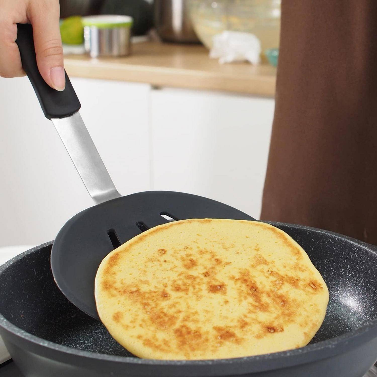 Hand, die mit einem Spatel einen goldenen Pfannkuchen in einer Pfanne auf einem Küchenherd wendet.