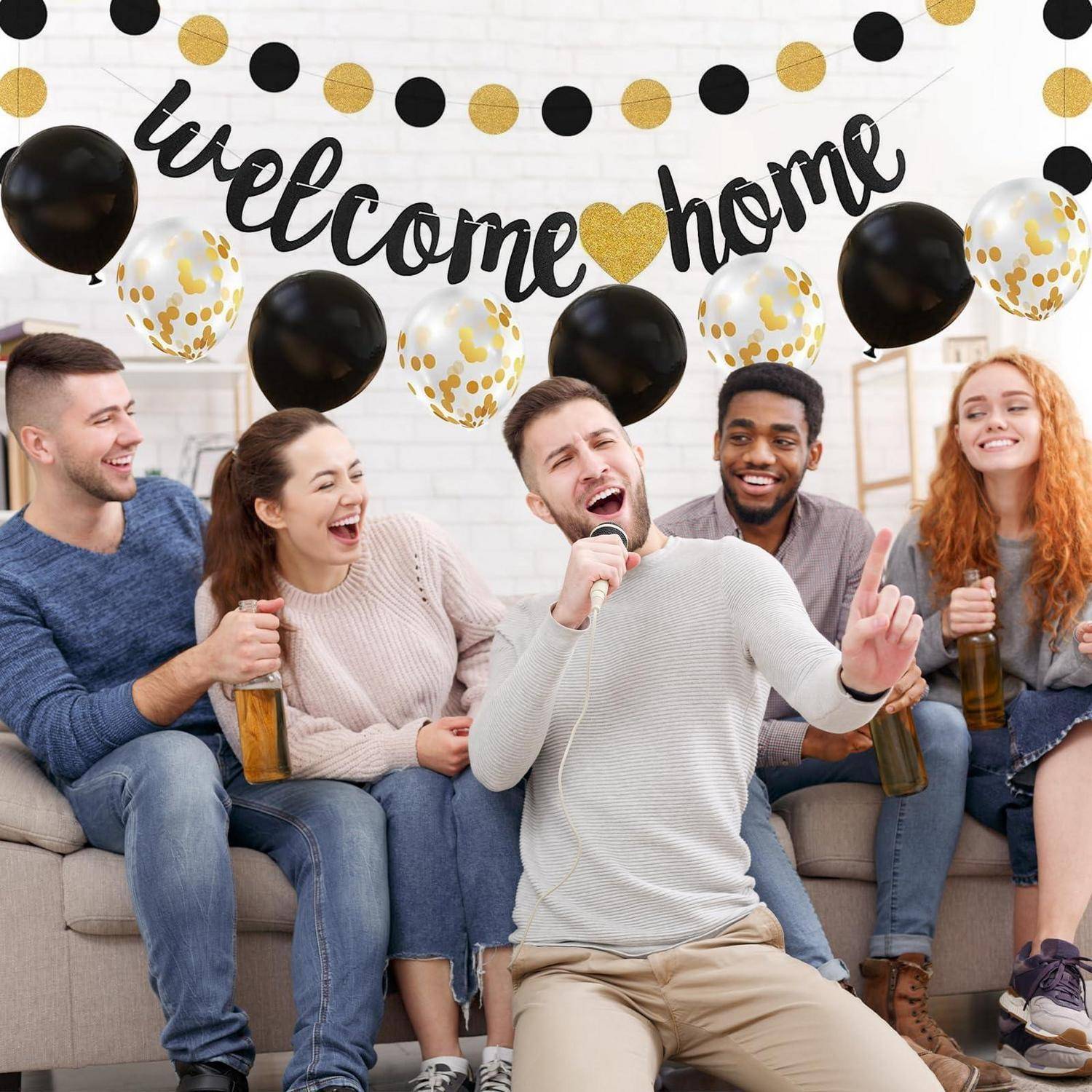Freunde auf einer Hausparty singen und lachen auf einer Couch unter einem "Willkommen zu Hause""-Banner mit schwarzen, goldenen und Konfetti-Ballons.