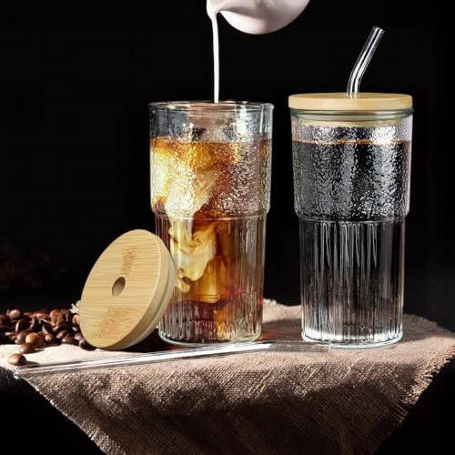 Zwei eisgekühlte Kaffegetränke in Glaspokalen mit Bambusdeckeln und Strohhalmen auf einem Tuch, wobei Milch in eines der Getränke gegossen wird.