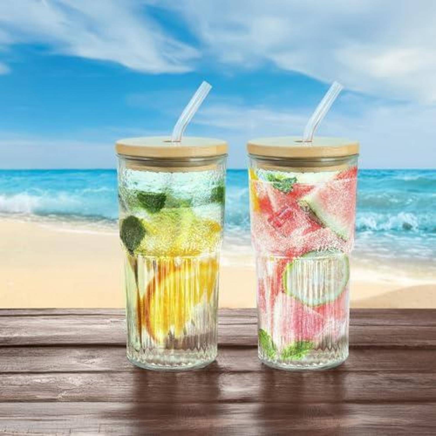 Zwei fruchtige Getränke in klaren Gläsern mit Strohhalmen stehen auf einem Holztisch, im Hintergrund sind ein Strand und das Meer zu sehen.
