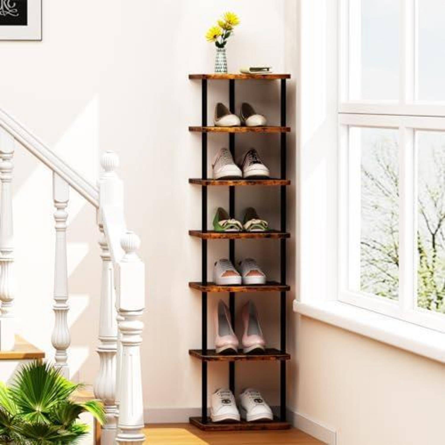Hohes, schmales Schuhregal neben einer Treppe und einem Fenster, das mehrere Paar Schuhe enthält, mit einer kleinen Vase mit gelben Blumen oben drauf.