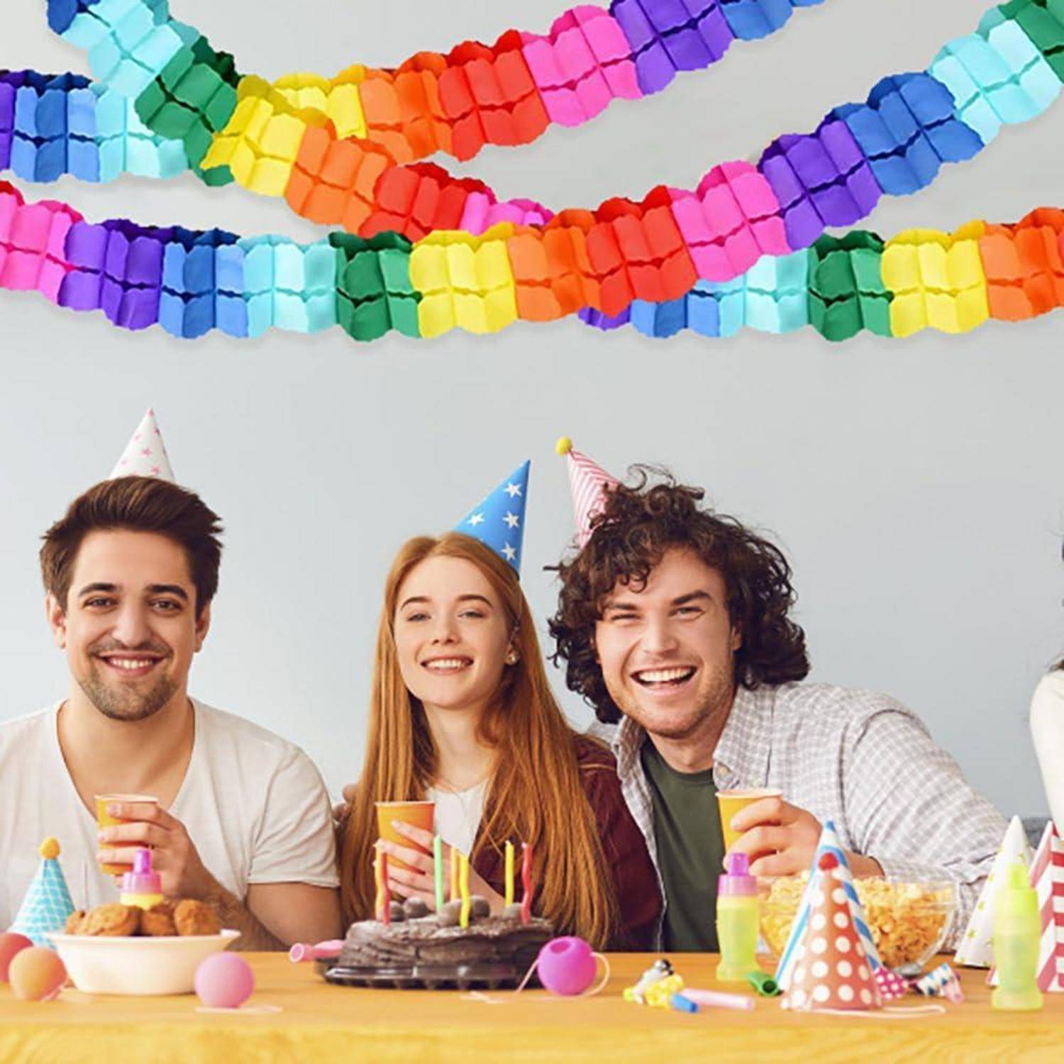 Drei lächelnde junge Erwachsene in Partyhüten feiern an einem Tisch mit Kuchen, Snacks und Getränken unter bunten Papiergirlanden.