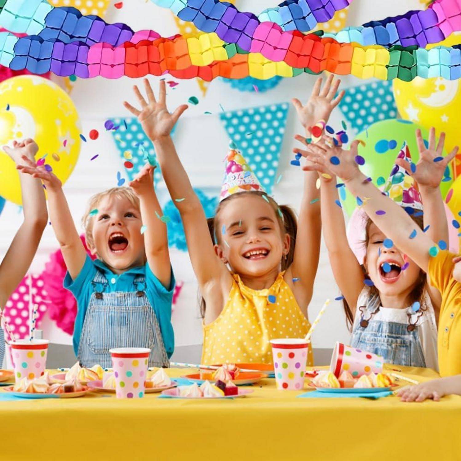 Drei Kinder auf einer farbenfrohen Geburtstagsfeier heben ihre Hände in die Luft und jubeln zwischen Konfetti, Luftballons, Girlanden und einem Kuchtentisch.