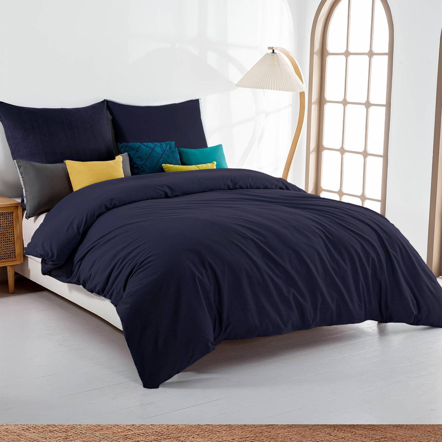 Ein sorgfältig gemachtes Bett mit dunkelblauer Bettwäsche und farbenfrohen Akzentkissen in einem hellen, minimalistischen Schlafzimmer, neben einer Stehleuchte und einem gewölbten Spiegel.