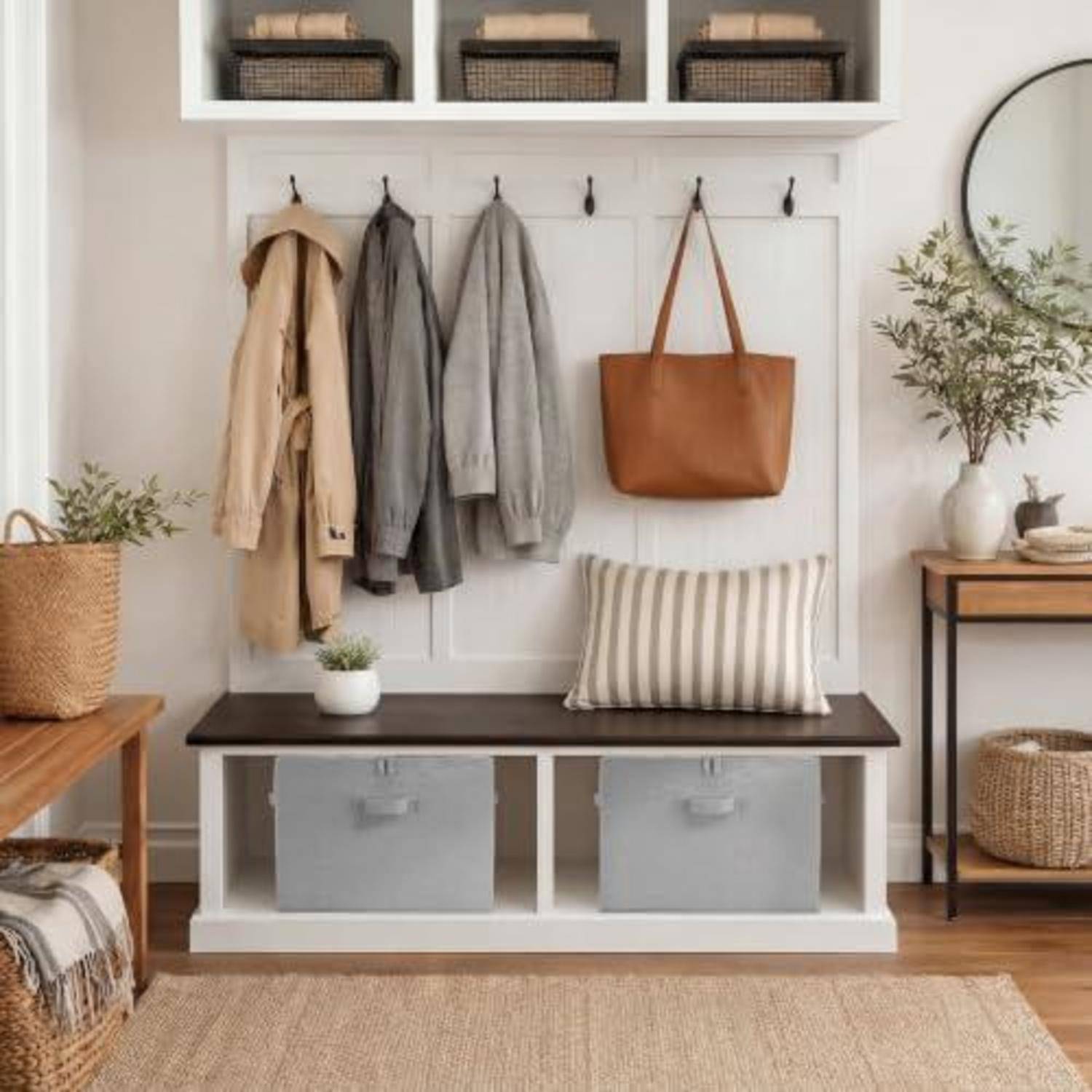 Eingangsbereich mit Garderobe mit Wandhaken, an denen Mäntel und eine beige Tragetasche hängen, eine Bank mit Aufbewahrungsbehältern und Kissen, geflochtene Körbe und ein Beistelltisch mit einer Pflanze.