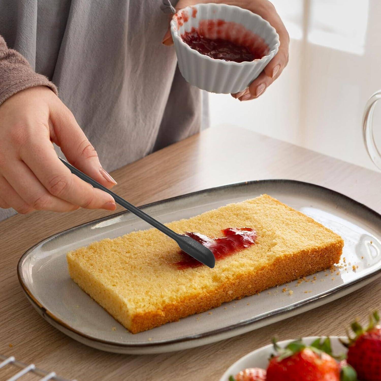 Eine Person verteilt rote Marmelade auf einen rechteckigen Schwammkuchen auf einem Tablett und hält dabei eine kleine Schüssel mit Marmelade; Erdbeeren sitzen in der Nähe.