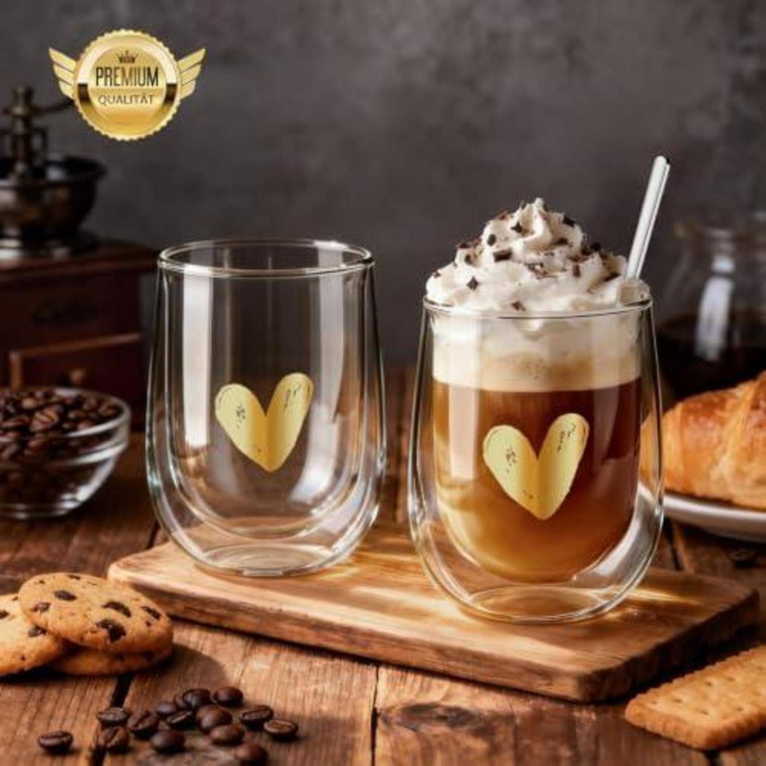 Zwei doppelwandige Glastassen auf einem Holzbrett, eine leer und eine gefüllt mit aufgeschäumtem Kaffee, oben mit Creme bedeckt; beide haben goldene Herzdesigns.