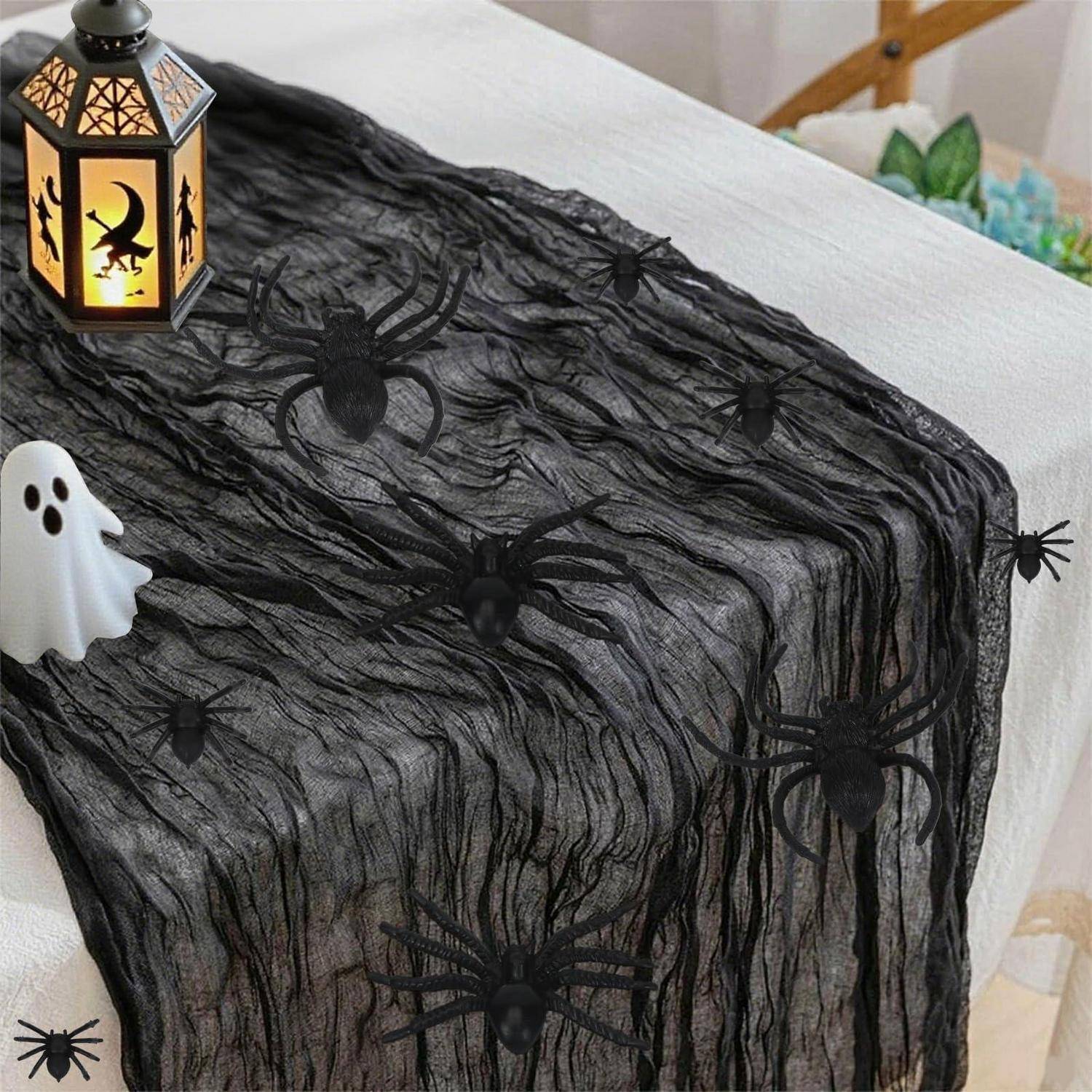 Halloween-Tischdekoration mit schwarzem, gaze-ähnlichem Netzstoff, Kunststoffspinnen, einer kleinen Geistfigur und einer leuchtenden Laterne auf einer weißen Oberfläche.