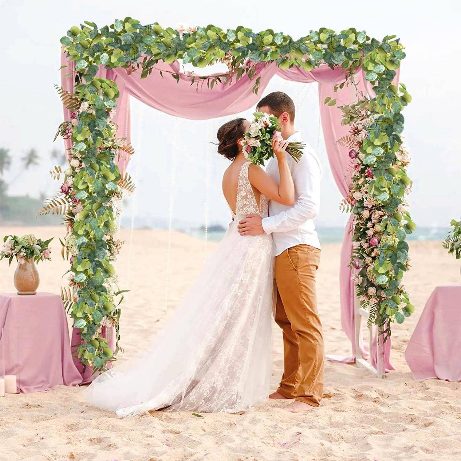 Braut und Bräutigam umarmen sich und küssen sich unter einem rosa drapiertem Blumenbogen bei einer Hochzeitszeremonie am Sandstrand.
