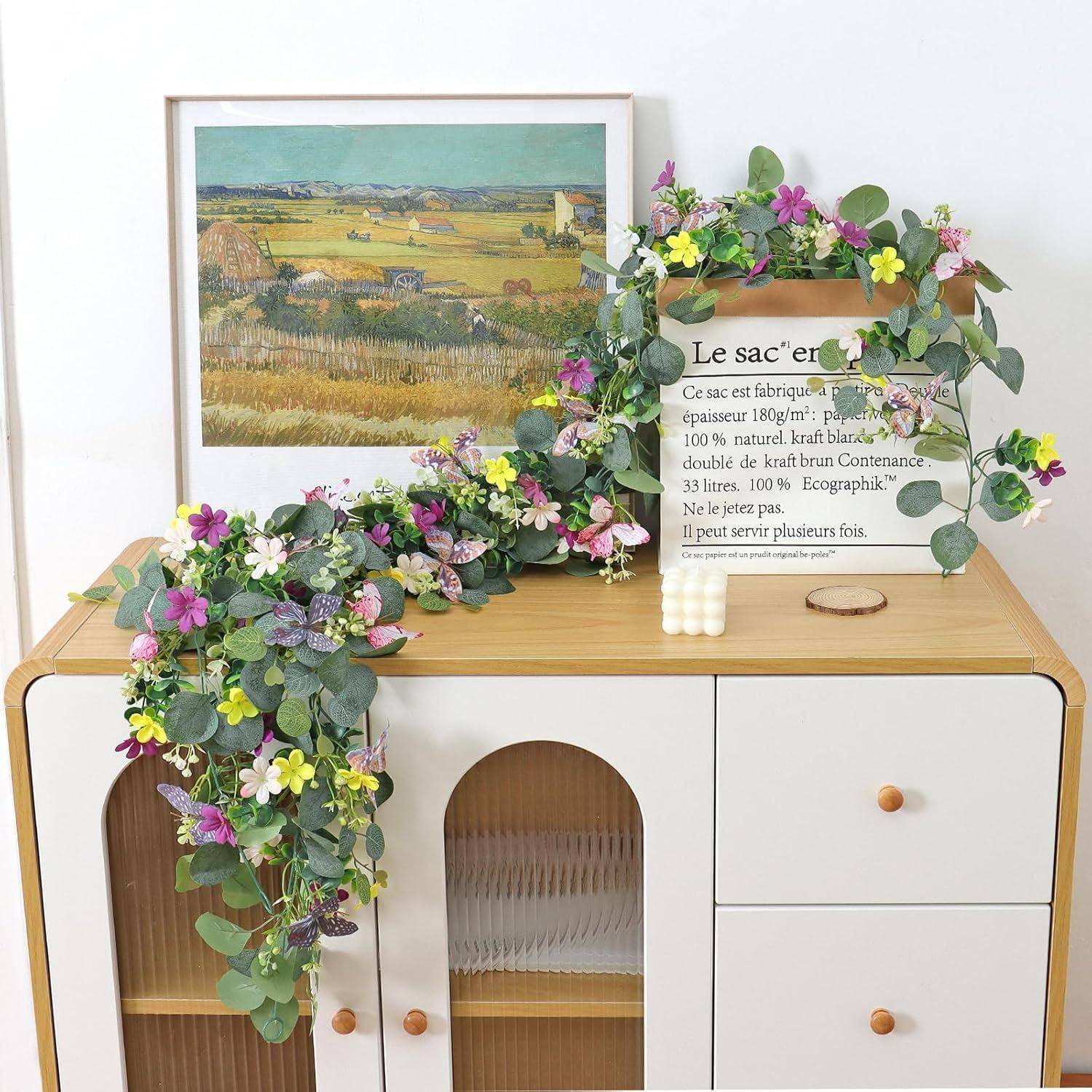Ein Holzschrank mit einem gerahmten Landschaftsdruck und einem dekorativen Schild, drapiert mit einer farbenfrohen Blüten- und Blattgirlande.