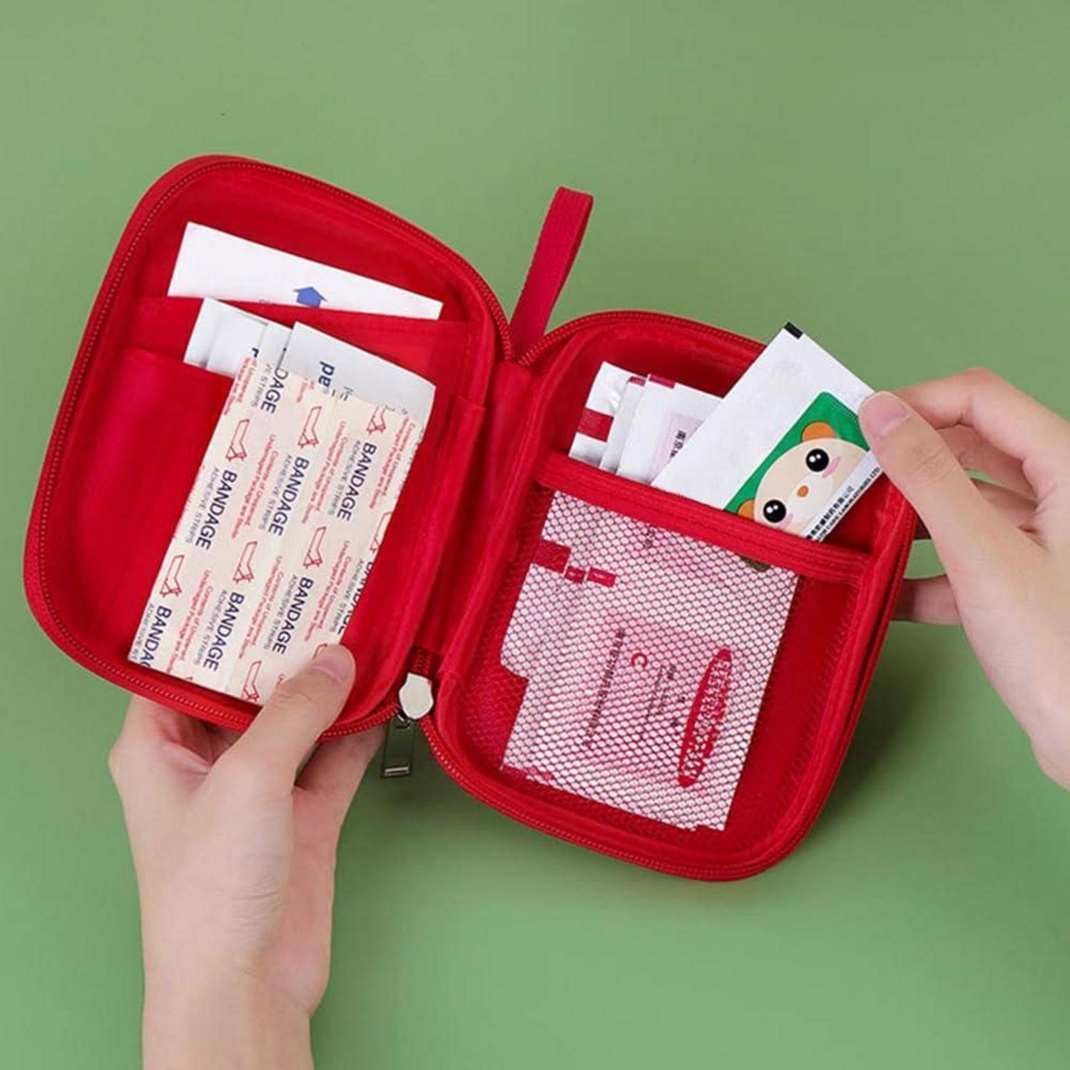 Offene rote Reißverschluss-Erste-Hilfe-Tasche mit Verbandsmaterial und kleinen medizinischen Hilfsmitteln, die von zwei Händen vor grünem Hintergrund geöffnet wird.