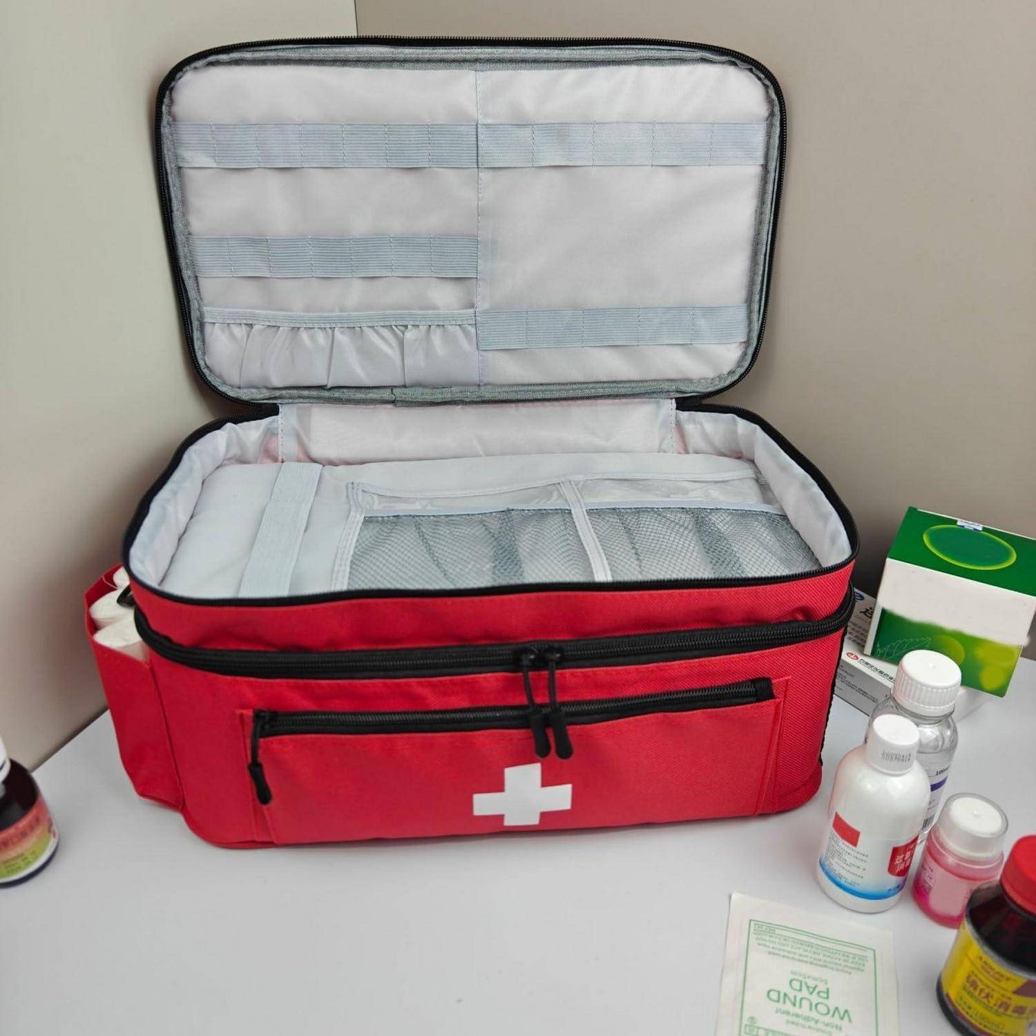 Offene rote Erste-Hilfe-Tasche mit organisierten Innentaschen, weißem Kreuz auf der Vorderseite und mehreren Medikamentenflaschen daneben auf einem Tisch.