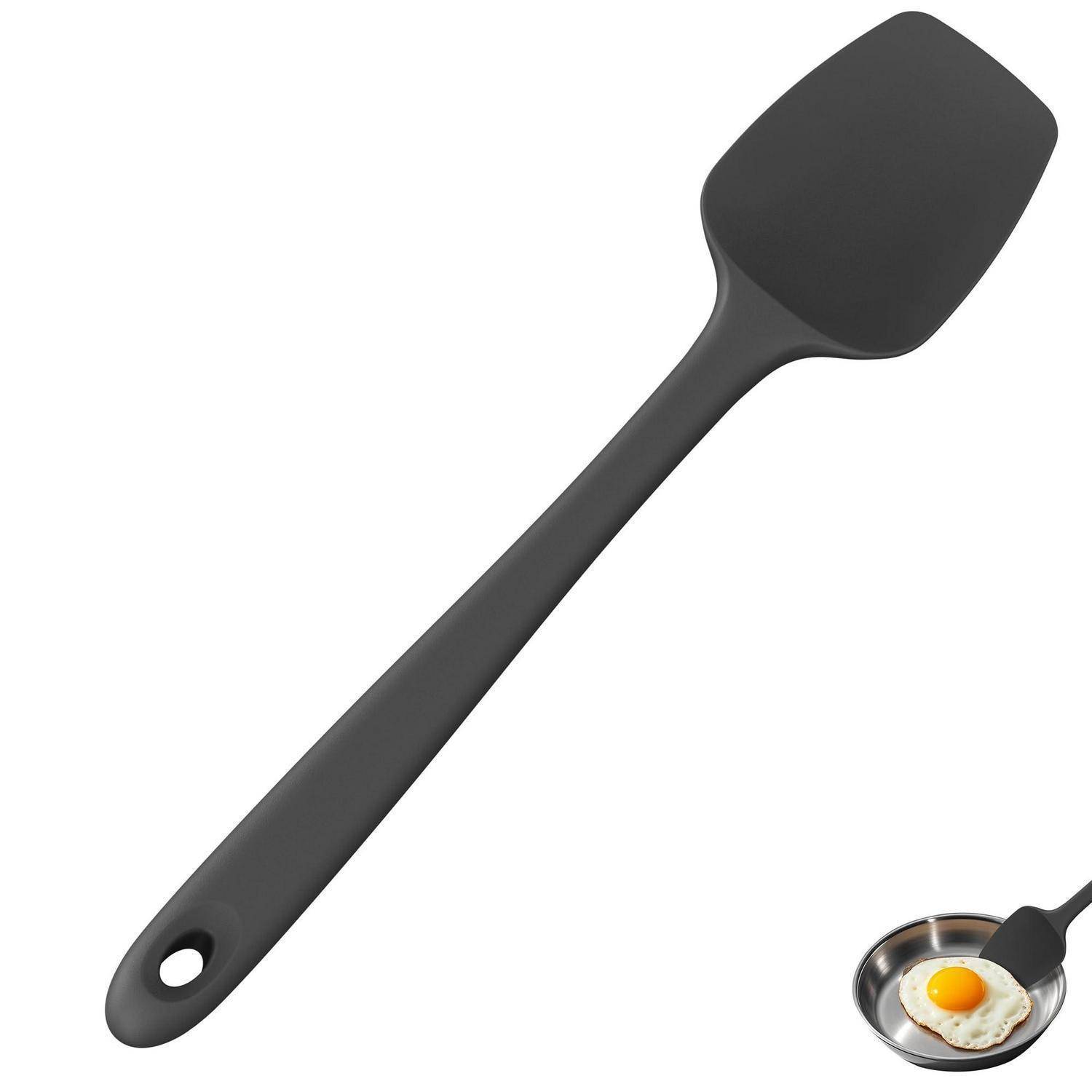 Schwarzer Silikonspatel mit Aufhängeloch, auf weißem Hintergrund neben einer kleinen Pfanne mit einem Spiegelei gezeigt, was die Verwendung in der Küche beim Kochen anzeigt.