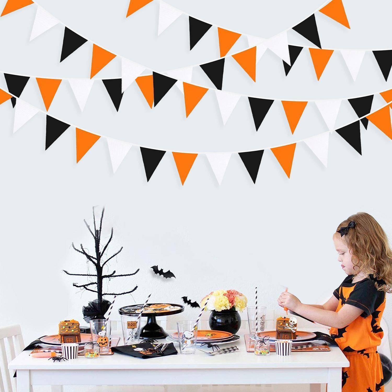 Kind in einem orange-schwarzen Kostüm dekoriert einen Halloweentisch mit Kuchen, Süßigkeiten, Fledermäusen und Wimpelketten auf weißem Hintergrund.